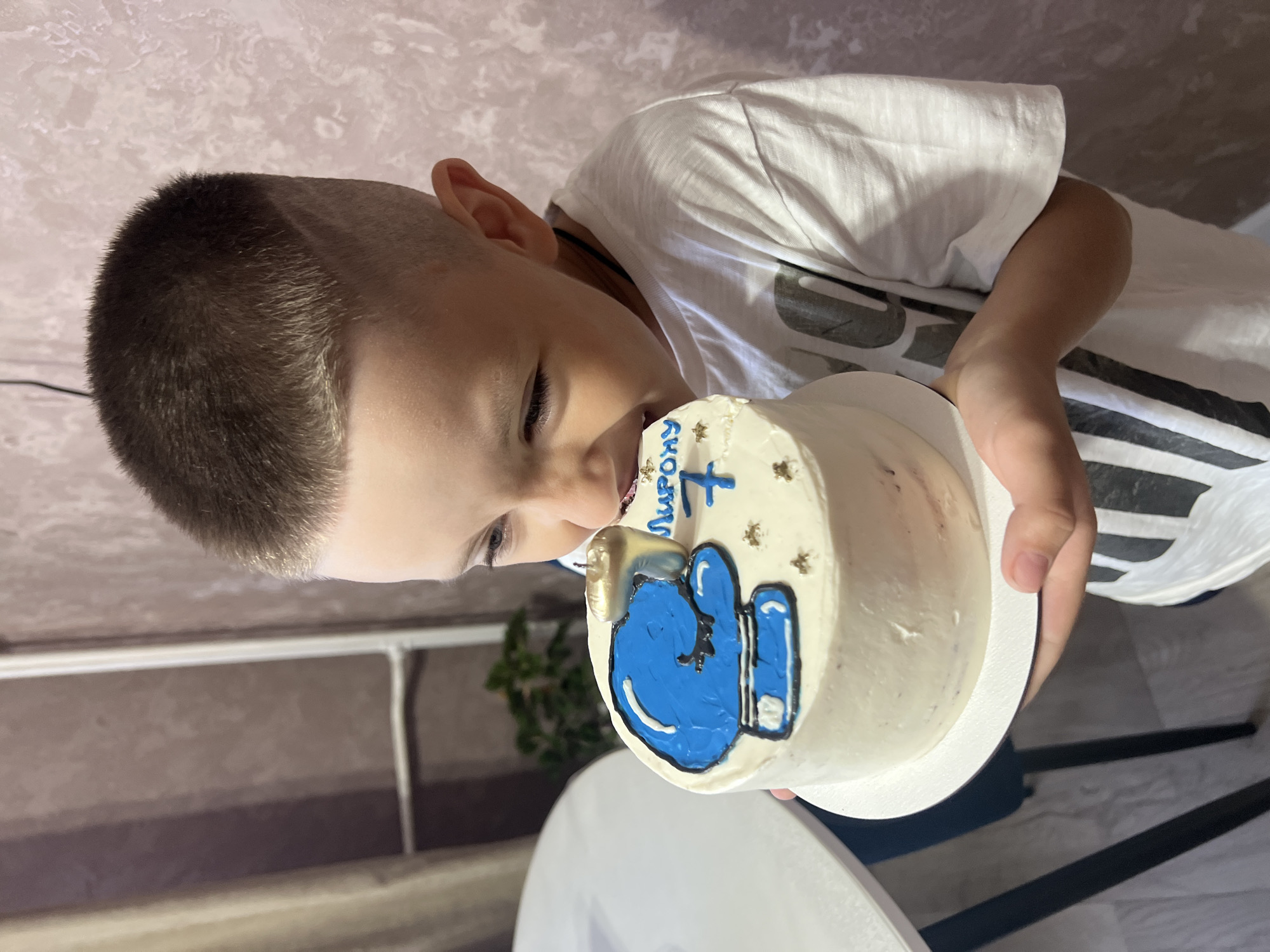 A young boy, approximately seven years old, is captured in a close-up shot as he leans in to bite a birthday cake. He is wearing a white t-shirt with dark stripes. The cake, round and white, is decorated with a blue image that resembles a character or emblem, and has the number "7" as a candle. The boy's face shows concentration and enjoyment as he interacts with the cake. The background is softly lit with a textured wall and some blurred elements of furniture, possibly in a home environment. The image appears to be taken in the city of Марковский, Russia.