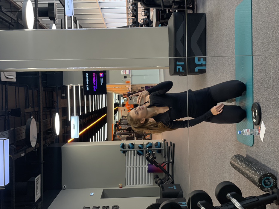 A young woman in black athletic wear takes a selfie in a gym mirror. She kneels on a turquoise yoga mat with a water bottle and weight nearby. Behind her are various exercise equipment, including dumbbells and medicine balls. The reflection shows a long gym with rows of exercise machines, and signs with text in a foreign language.