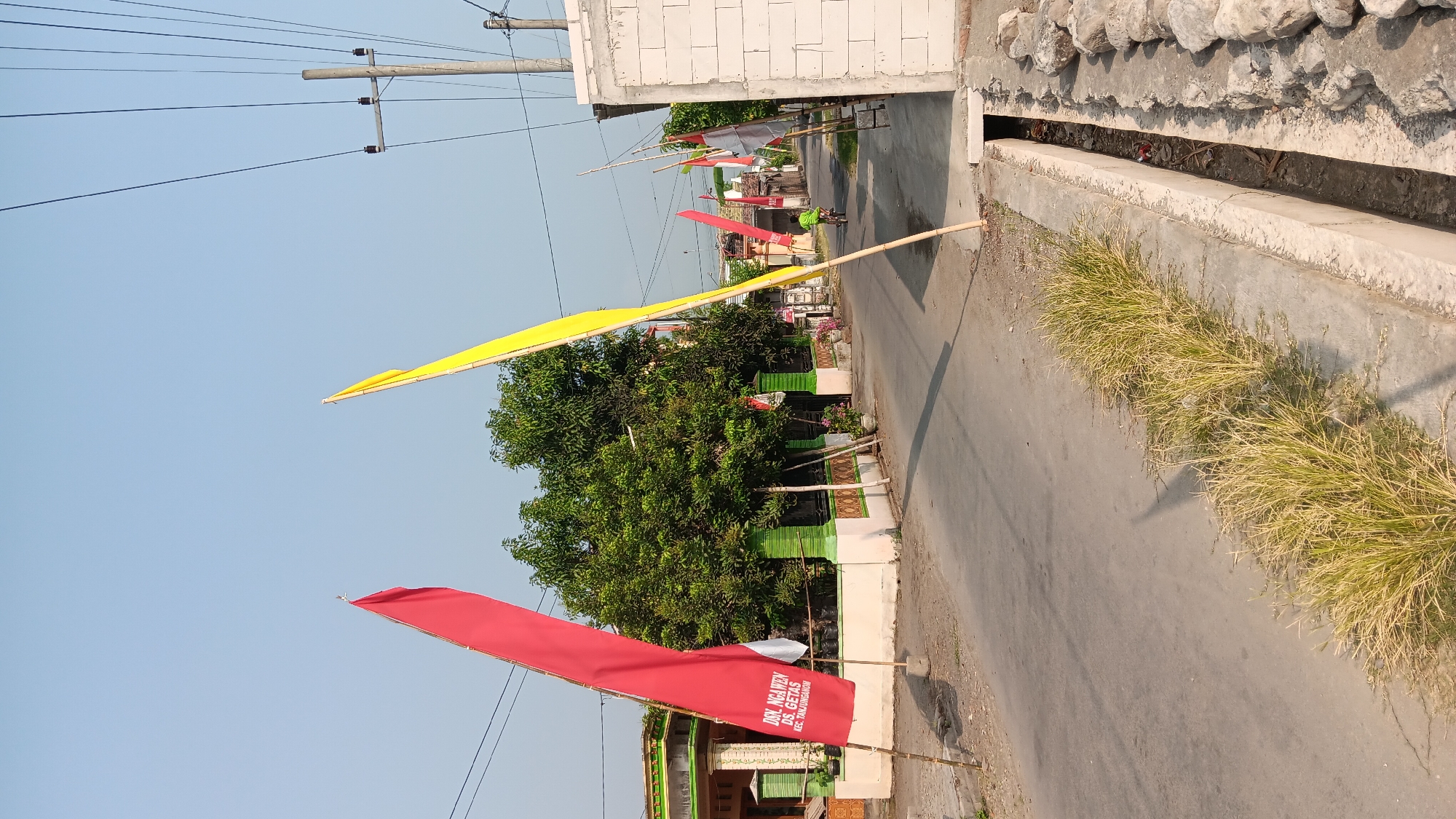 This image depicts a street scene in Sonobekel, Indonesia, likely a village or rural area given the presence of traditional houses and unpaved sections of the road. Two large flags, one red and one yellow, are prominently displayed on wooden poles. The red flag, in the foreground, has text written on it, appearing to be a place name. A person on a bicycle is visible in the middle ground, riding down the street. More flags, including some with red and white stripes, are scattered further down the road. The environment includes houses with green trim, mature trees, a concrete drainage ditch, and utility poles with wires. The sky is clear and blue, suggesting it is daytime and likely fair weather. The overall impression is one of a quiet residential street decorated for an event or celebration.