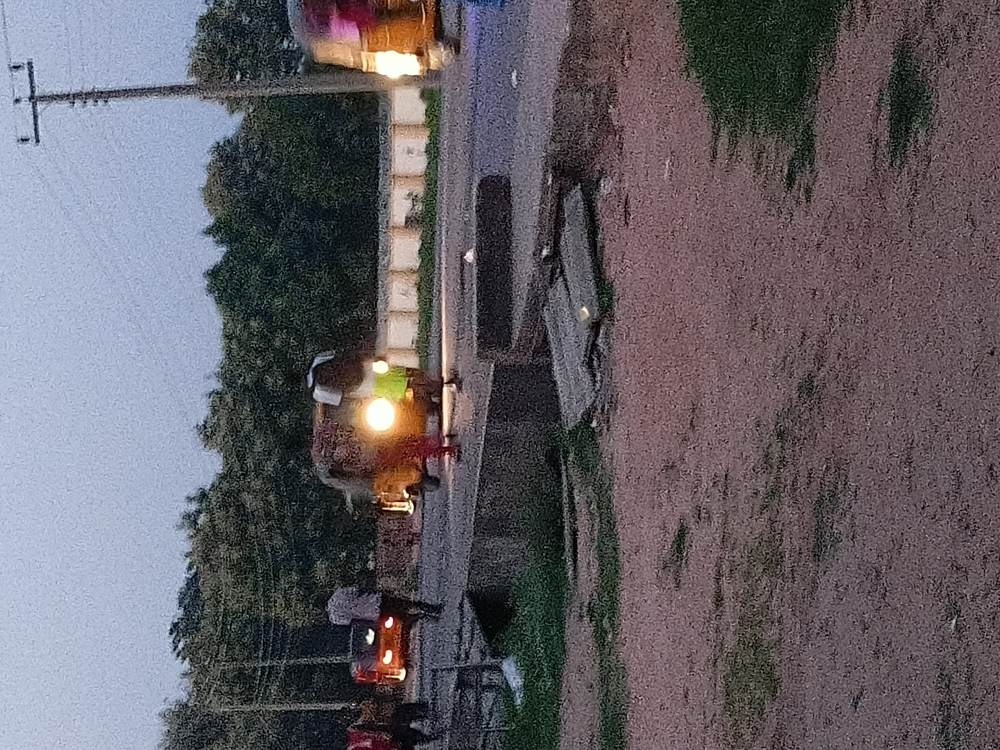 The image depicts an outdoor scene in Jalingo, Nigeria, at dusk. A paved road runs horizontally through the frame, bordered by a low concrete structure on the right and a dirt area with sparse vegetation on the left. A wooden utility pole with crossbars stands prominently on the right side of the image, indicating it's a built-up area.
In the middle ground, a person in a light-colored shirt walks away from the viewer towards the left, beside a small, orange vehicle with its taillights illuminated. To the right of this vehicle, a three-wheeled auto-rickshaw, also known as a "Keke," is visible, its headlights glowing brightly. The Keke appears to be in motion or stopped with its engine running. Another auto-rickshaw, partially obscured and with its headlights on, is further to the right, and its front is largely out of frame.
In the background, dense green trees form a backdrop, with a low, light-colored wall or fence running along the edge of the road. The sky is overcast and appears to be a light gray, typical of twilight. The ground in the foreground is rough and uneven, composed of dirt and small stones, with patches of green grass. The overall impression is a street scene in a Nigerian city as evening descends, with vehicular traffic and pedestrian activity.
