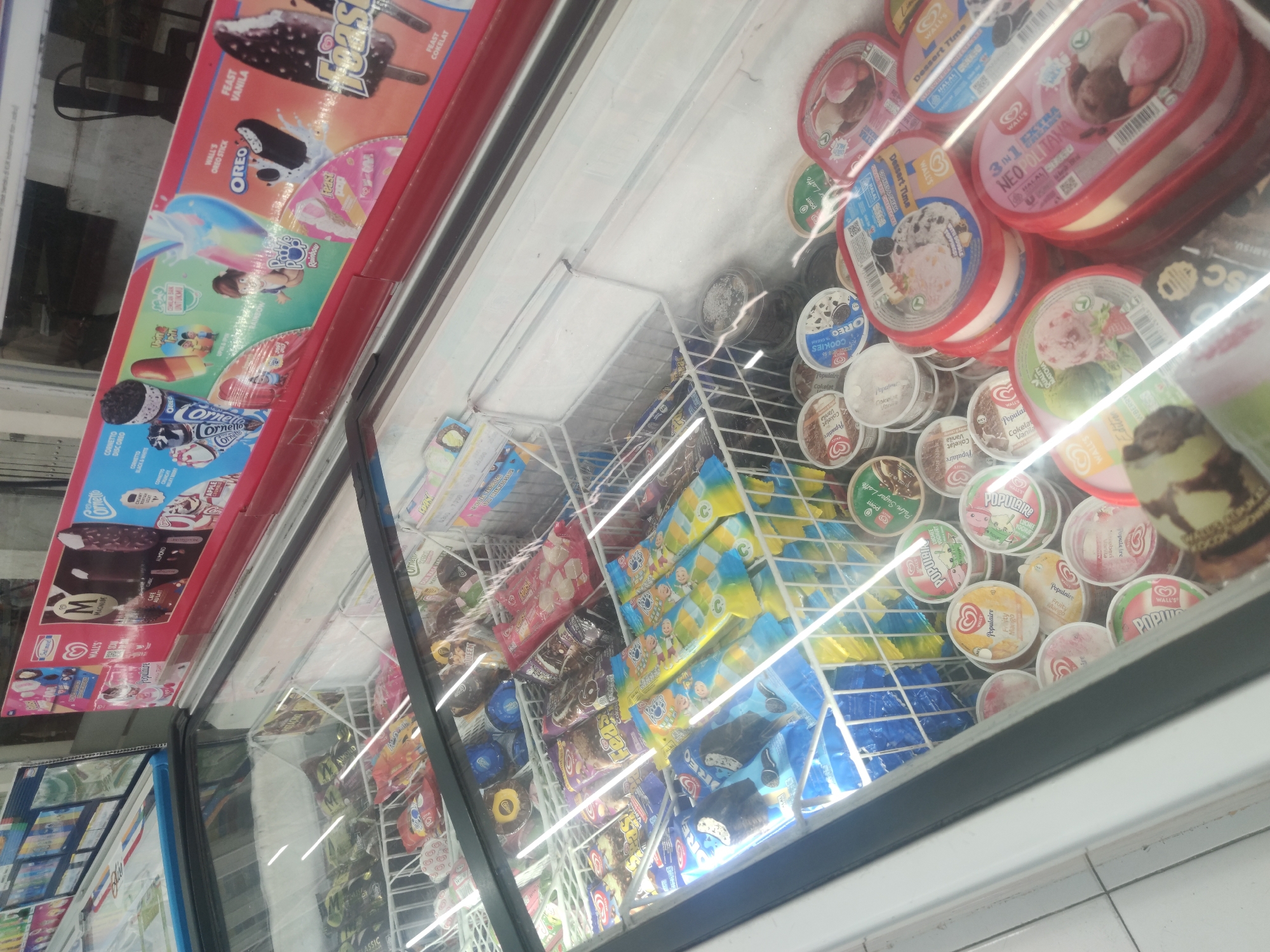 This image captures a refrigerated display case at a convenience store, likely located in Surabaya, Indonesia. The case is filled with a variety of frozen treats, primarily ice cream products from the Wall's brand. Prominent items include packaged ice cream bars like Cornetto and Feast, as well as tubs of ice cream with flavors such as Oreo, Populaire, and various fruit options like mango and strawberry. The packaging indicates a range of flavors and types, with some featuring cartoon characters suggesting they are targeted towards children. The lighting within the display case is bright, illuminating the products. The background shows glimpses of shelving and other store fixtures, consistent with a retail environment.