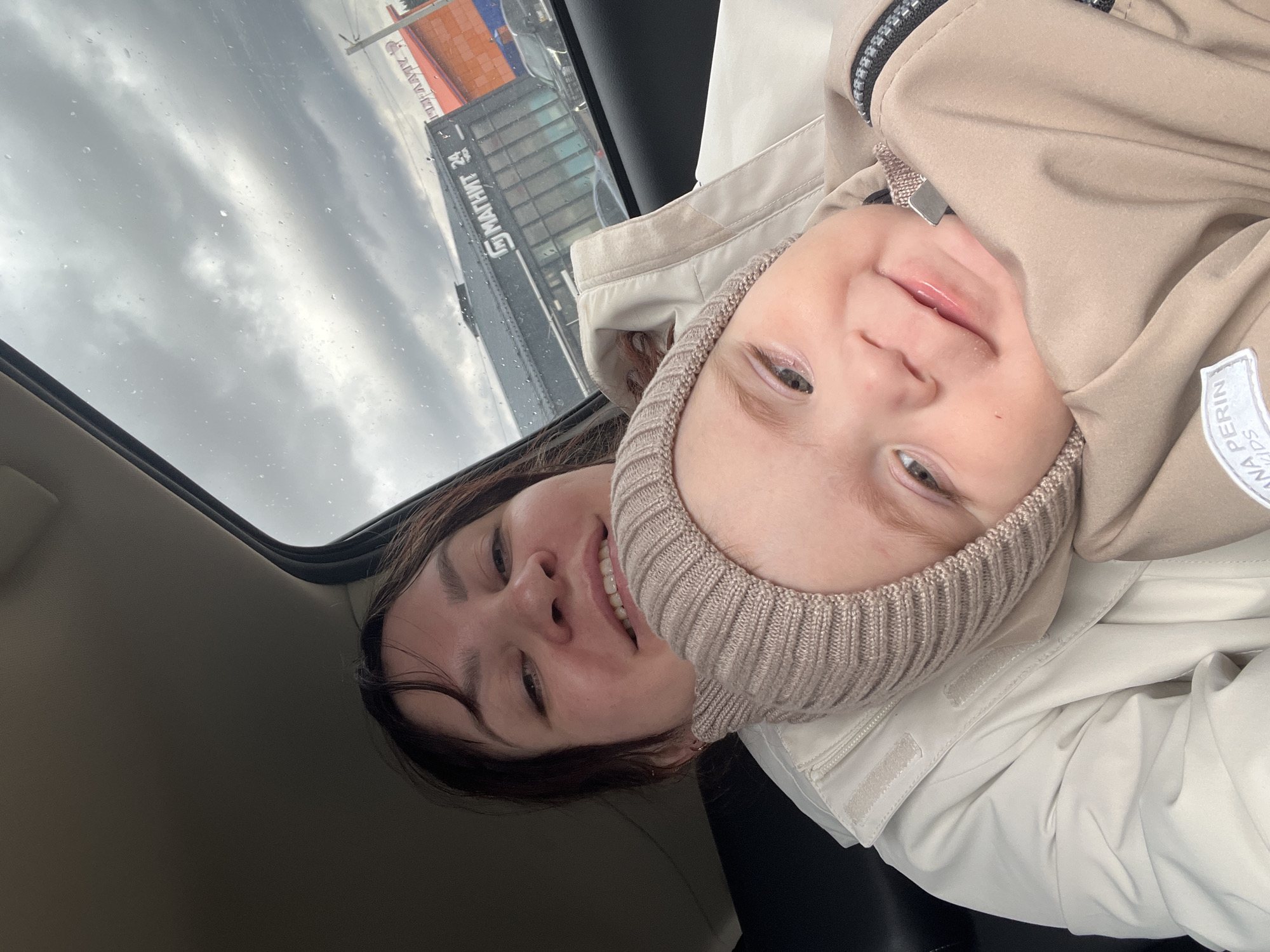 A selfie-style photograph captures a woman and a young child inside a vehicle, likely in Protasy, Russia. The image is taken from a low angle, looking upwards through the car's sunroof or window. Both individuals are smiling broadly, looking directly at the camera.
The woman, with dark hair, is seen smiling, revealing her teeth. She wears a light-colored jacket over a dark top. The child, likely a baby or toddler, wears a light beige knitted hat and a matching beige jacket. The child's jacket features a white patch with "ANNA PERIN KIDS" visible on it.
Through the vehicle's sunroof, an overcast, cloudy sky is visible, with raindrops evident on the glass, suggesting wet weather. Beyond the vehicle, reflections of buildings are visible. One prominent reflection shows a building with the Cyrillic text "МАТНИМ 24" which is a reversed reflection of "МАГНИТ 24," indicating a 24-hour Magnit supermarket, a major Russian retail chain. Another building with an orange-red roof is also faintly visible. The scene conveys a cheerful and intimate moment despite the gloomy outdoor conditions.