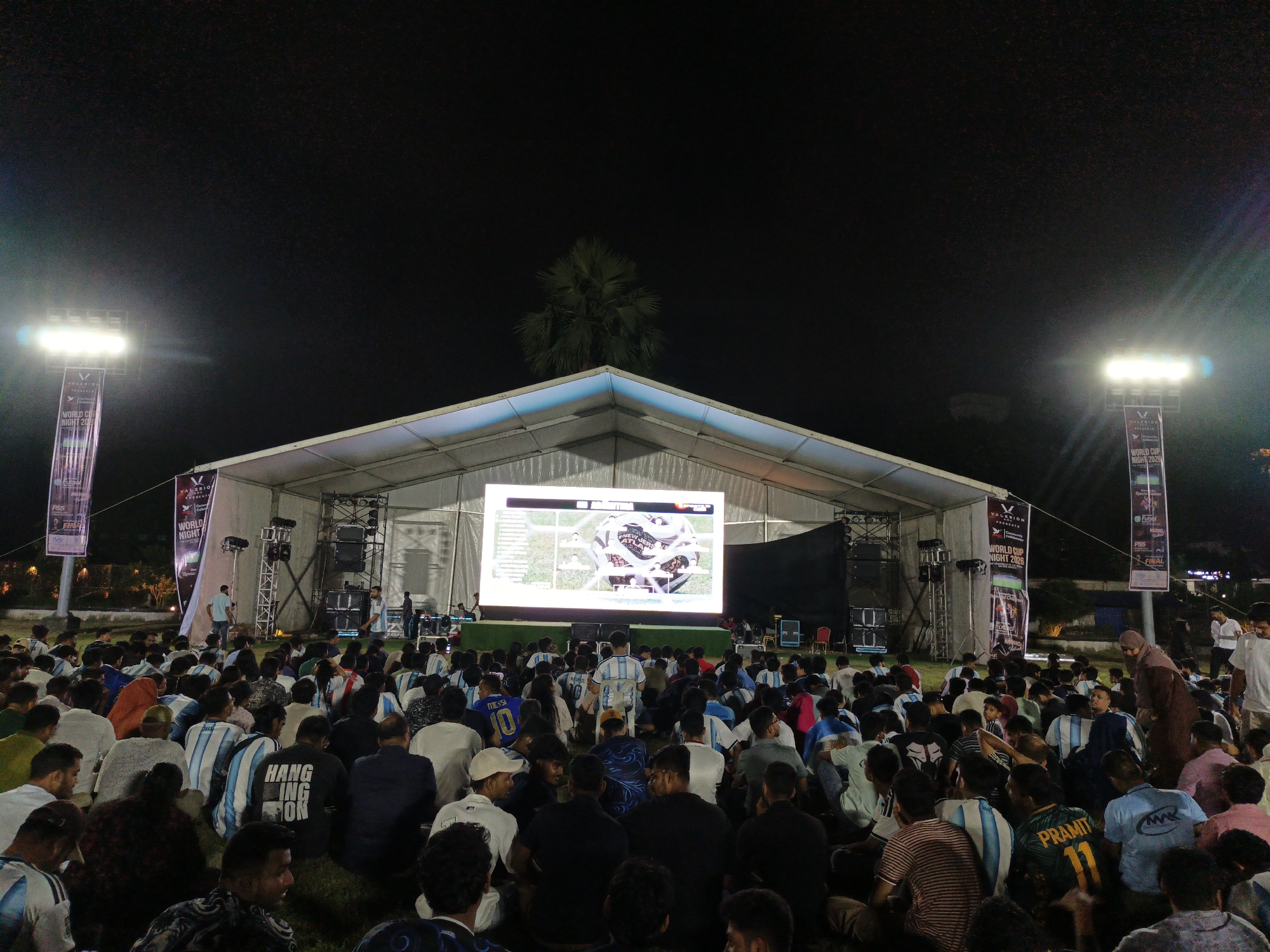 An outdoor event is taking place at night under a large tent structure in Dhaka, Bangladesh. A large LED screen displays a football game, with a close-up of a football in the center. A crowd of people, many wearing football jerseys, are seated on the ground watching the screen. Two tall banners with the text "VALERION PRESENTS WORLD CUP NIGHT 2026" are visible on either side of the tent. Stage lighting illuminates the area.