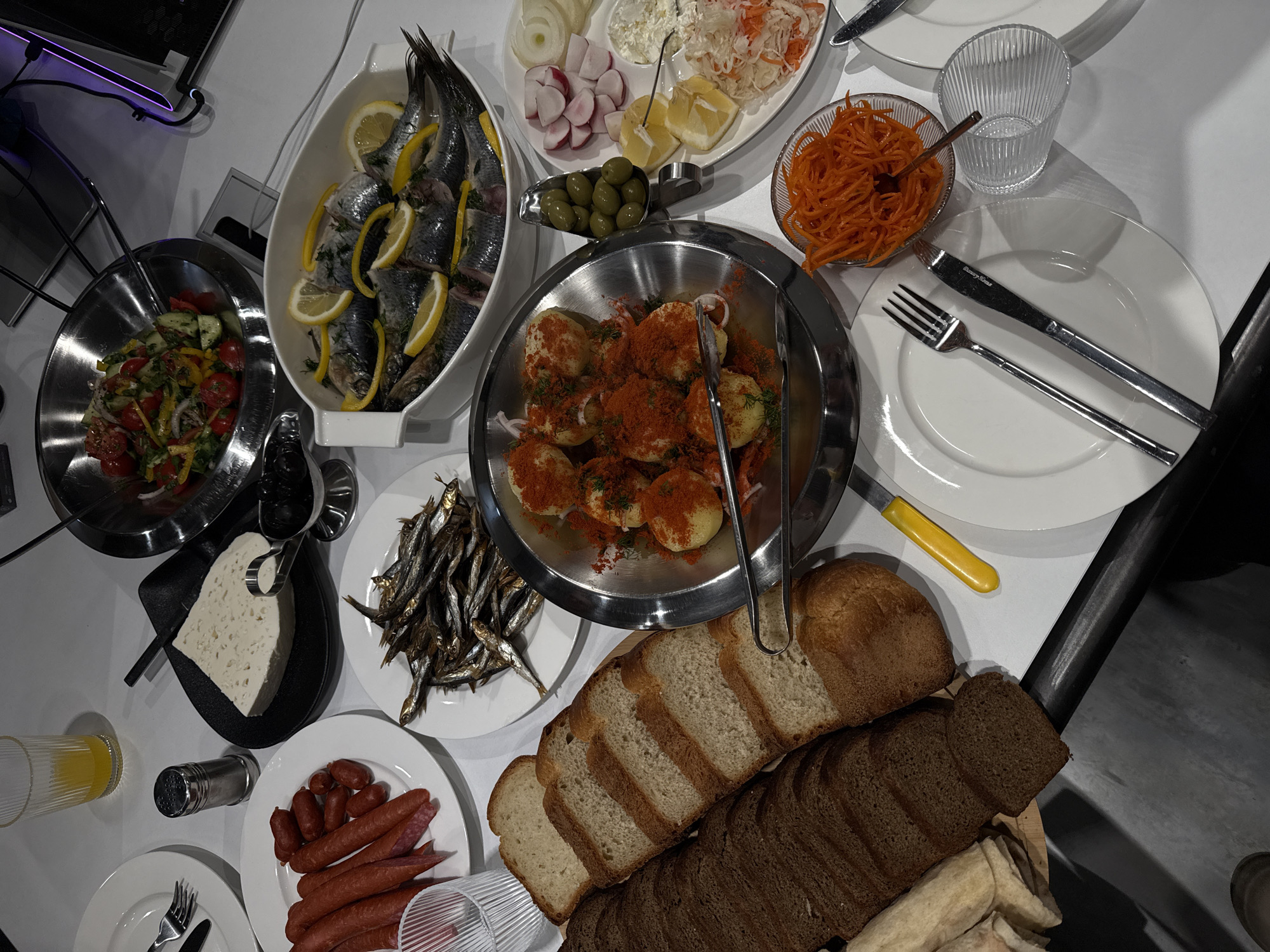 The image depicts a table laden with food, seemingly a spread for a meal. There is no clear indication of the exact location within Yerevan, Armenia, or whether the event is public or private. The focal point is the diverse assortment of dishes. A white platter contains a large fish, possibly herring or mackerel, garnished with lemon slices. Other dishes include various salads, one with sliced tomatoes and peppers, another with shredded carrots, and a platter with sliced radishes, onions, and a white spread. Small, fried fish lie on a white plate. Boiled potatoes sprinkled with paprika are presented in a metal bowl. A selection of breads, including white and dark rye, is arranged near the edge of the table. Sausages are present on a white plate. Additionally, a glass of yellow liquid sits beside a salt shaker. Silverware and white plates are set around the table, indicating that the food is ready to be served and eaten. There are no visible people in the frame, so there are no visible actions or interactions. The environment is a well-lit indoor setting.
