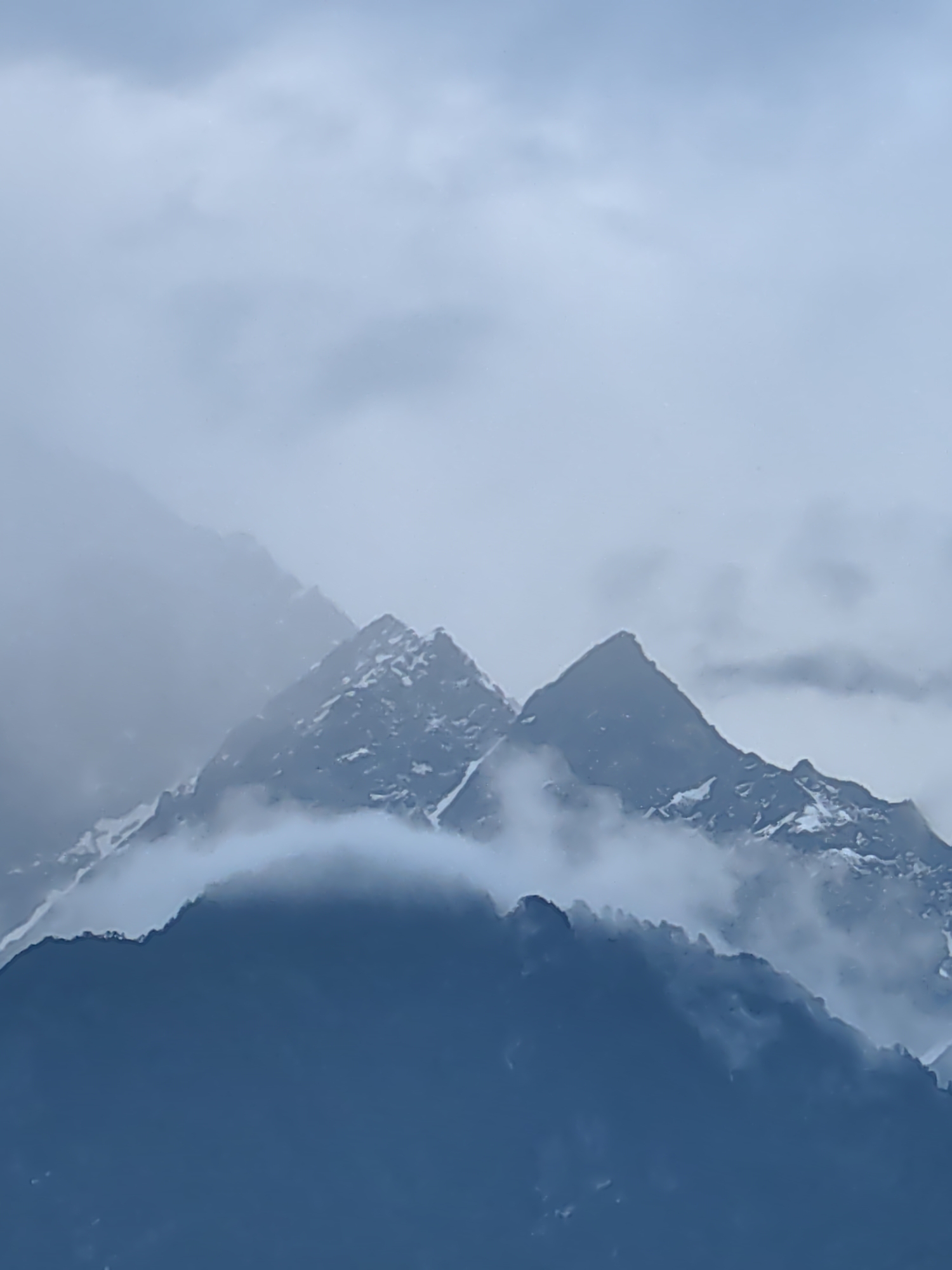 The image depicts a dramatic, cloudy mountain landscape with snow-capped peaks in Vashisht, India. The foreground is dominated by a dark, silhouetted mountain ridge, likely a lower elevation. Above this, the rugged, snow-dusted summits of taller mountains emerge from a thick blanket of mist and clouds, creating a sense of depth and mystery. The lighting is diffused and overcast, typical of a cloudy or misty day, with the sky filled with soft, grayish-blue clouds. The overall scene is serene and imposing, showcasing the raw beauty of the Himalayan terrain.