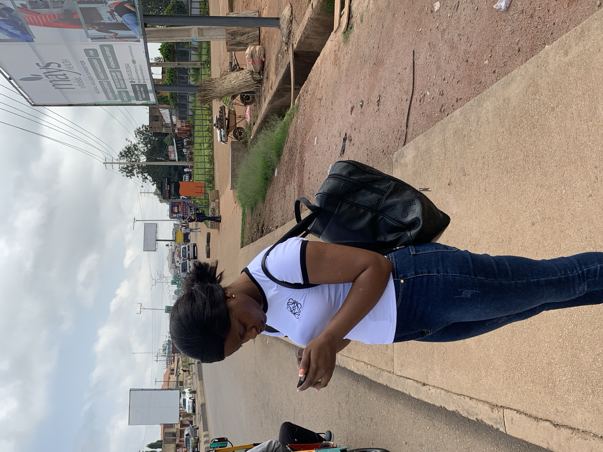 The image shows a young woman standing on a sidewalk in Gura Top, Nigeria, looking down at her phone. She is wearing a white t-shirt with black trim and dark blue jeans. A black backpack with a patterned logo is slung over her left shoulder. The background reveals a busy street scene with cars, billboards, and some greenery. The weather appears to be overcast. One billboard in the background displays text related to "mays wholistic HEALTH" and various wellness support services, along with a phone number and website. The overall impression is a candid moment of someone on their phone in an urban, possibly commercial area.