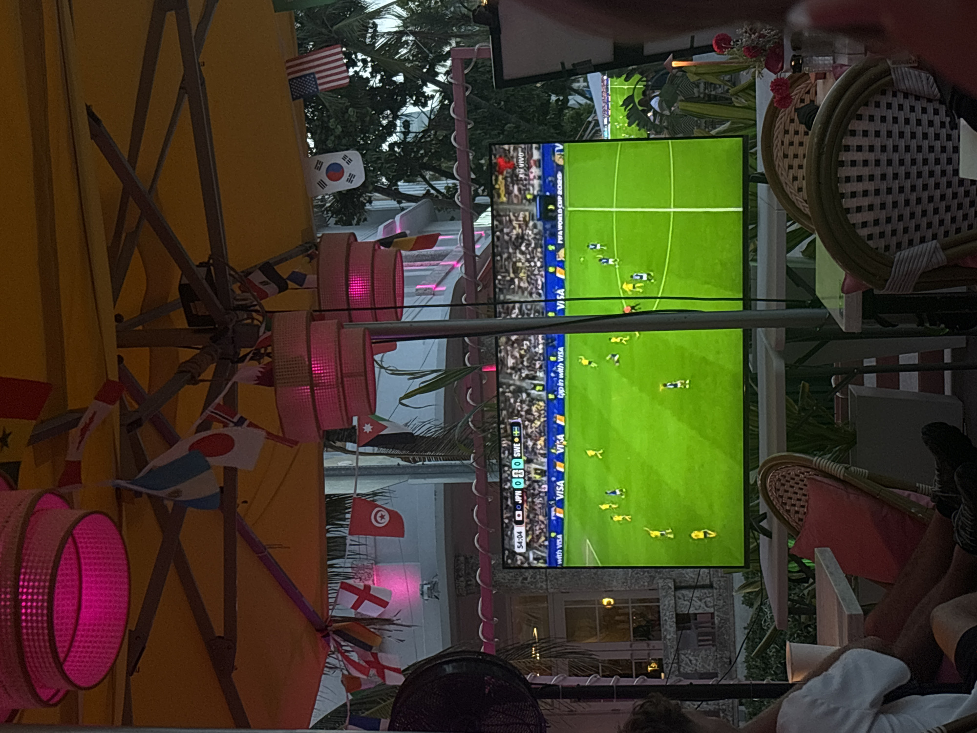 This image captures a lively outdoor scene, likely in Miami Beach, where people are gathered to watch a soccer match on a large screen. The setting appears to be a vibrant open-air venue, possibly a bar or restaurant, adorned with numerous national flags, suggesting a festive atmosphere related to an international sporting event like the FIFA World Cup.
The main subjects are the viewers, partially visible in the foreground, relaxing and focused on the game. We see legs and torsos of people seated on chairs and potentially on the ground. The chairs themselves are of a woven, outdoor style, adding to the casual, tropical ambiance. Hanging decorations, including pink, circular light fixtures and strings of small flags, contribute to the celebratory mood.
The screen prominently displays a soccer game in progress, with green turf and players in yellow and blue uniforms. Text on the screen indicates the match details: "54:04 JPN 0 0 SWE," suggesting the game is in its second half with Japan and Sweden tied. Other visible text includes "FIFA WORLD CUP 2020" and "VISA" advertisements, confirming the event and sponsorship.
The lighting suggests it might be late afternoon or early evening, as the outdoor lighting is illuminated, and the sky is not completely dark. The presence of palm trees in the background hints at a warm-weather location, consistent with Miami Beach. The overall impression is one of shared enjoyment and communal engagement with the sporting event, set against a visually rich and festive backdrop.