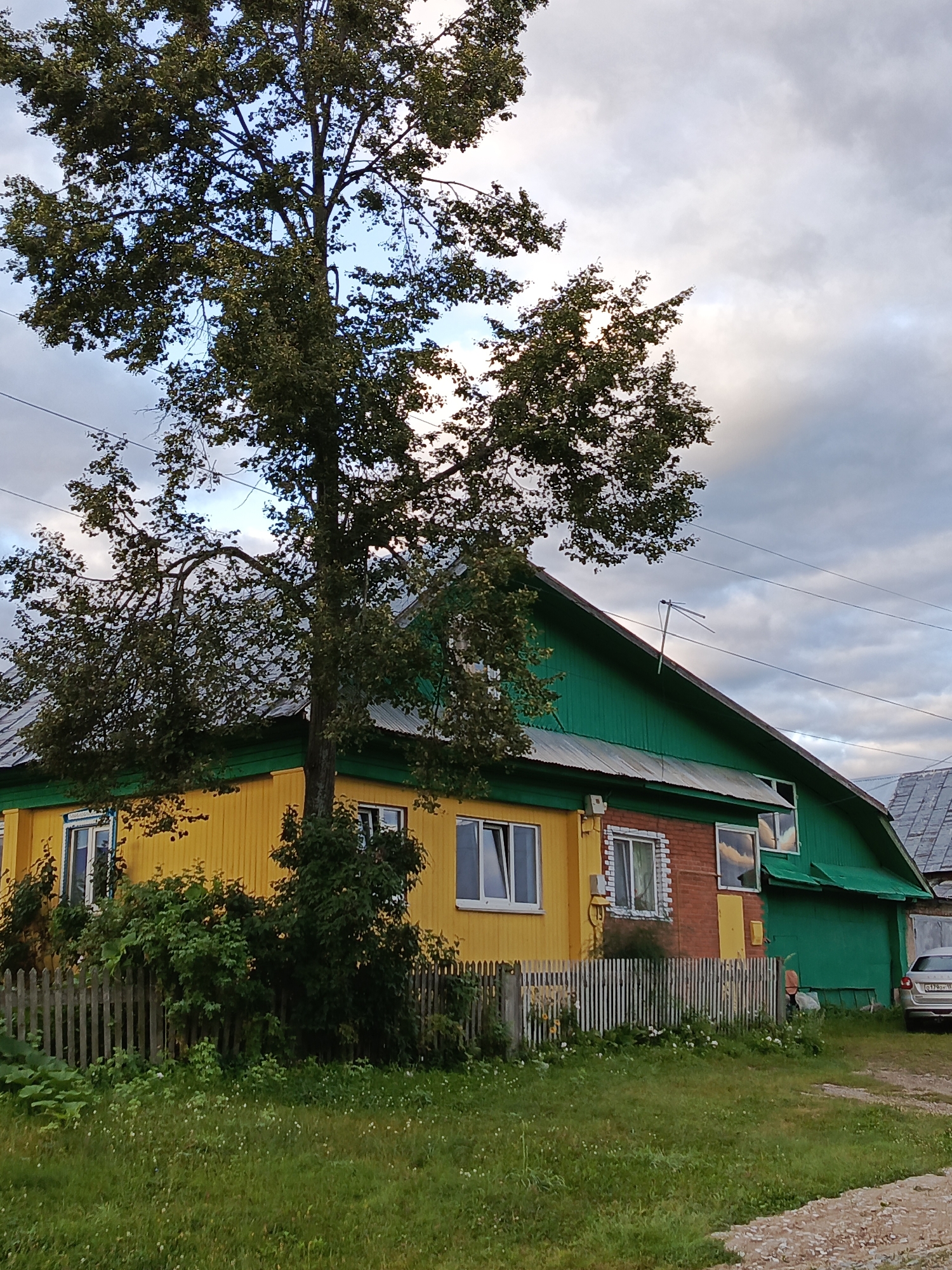 This is a medium shot of a yellow and green house with a large tree in front of it, taken in Chaikovsky, Russia. The house is two stories and has a sloped, green roof. The lower half of the house is yellow, with white-trimmed windows. The upper half is green, with some brick detailing and more white-trimmed windows. A white wooden fence runs along the front of the property. The tree in front of the house is tall and leafy, with some branches arching over the roof. The sky is cloudy and the lighting suggests it is late afternoon or early evening. In the background, a portion of another house and a car are visible. The ground in front of the house is covered in green grass with small white flowers.