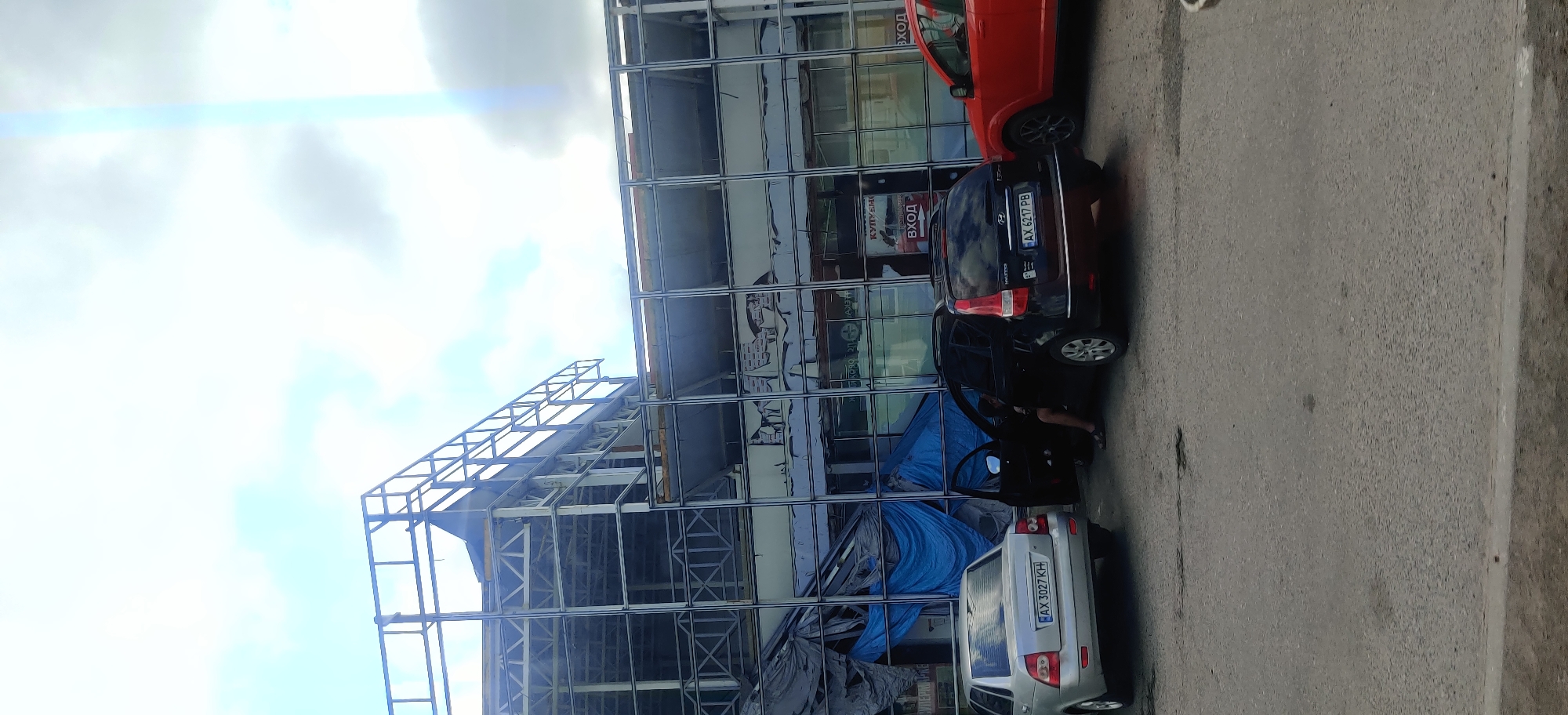 The image depicts a street scene in Valky, Ukraine, with a partially constructed or damaged building in the background. The building appears to be a commercial structure with large, reflective windows, some of which are covered with blue tarps. A metal frame structure is visible extending above the main building. In the foreground, three cars are parked on an asphalt road. A silver sedan is on the left, a black hatchback is in the center with its driver's door open and a person partially visible inside, and a red SUV is on the right. The road shows signs of wear, with some cracks and potholes. The sky is cloudy with a bright lens flare visible towards the upper right.