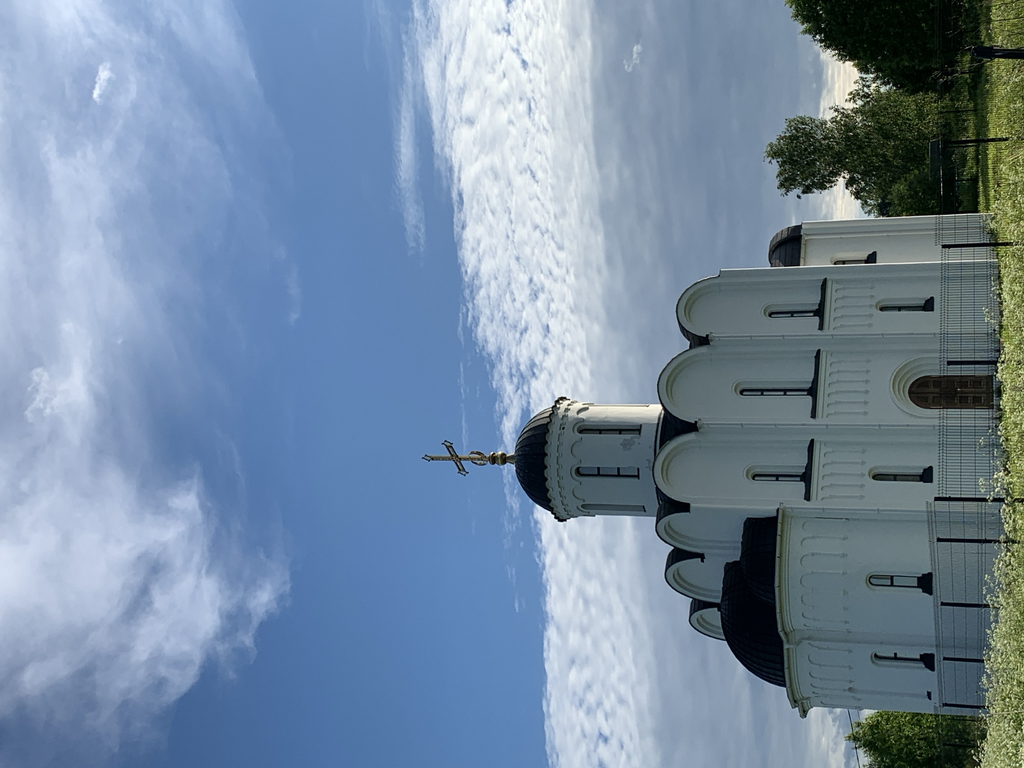 The image shows the exterior of a white church with multiple domes, one of which is topped with a golden cross. The church is situated in a green, grassy area with trees in the background. A metal fence runs along the side of the building. The sky is blue with scattered white clouds. The image is taken from a low angle, looking up at the church and sky. The time of day appears to be daytime, and the weather seems pleasant with clear skies and light clouds. The setting is likely a rural or suburban area given the presence of greenery and the relatively modest fence. There is no visible text in the image.