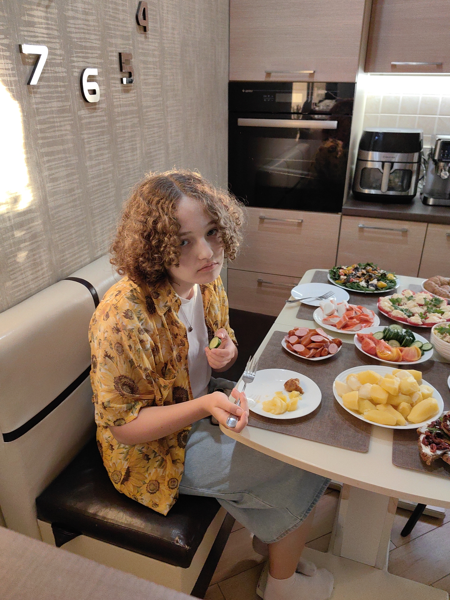 A young person with curly brown hair sits at a table laden with food in a kitchen. They are wearing a yellow floral shirt and a light blue skirt, and are holding a fork and a slice of cucumber. The table features several plates of food, including sliced sausages, crab sticks, salad, and potatoes. In the background, a kitchen oven and various appliances are visible. The wall behind the person has decorative numbers resembling a clock face. The scene appears to be a domestic setting, possibly a home in the city of Stowbtsy, Belarus, given the context provided.