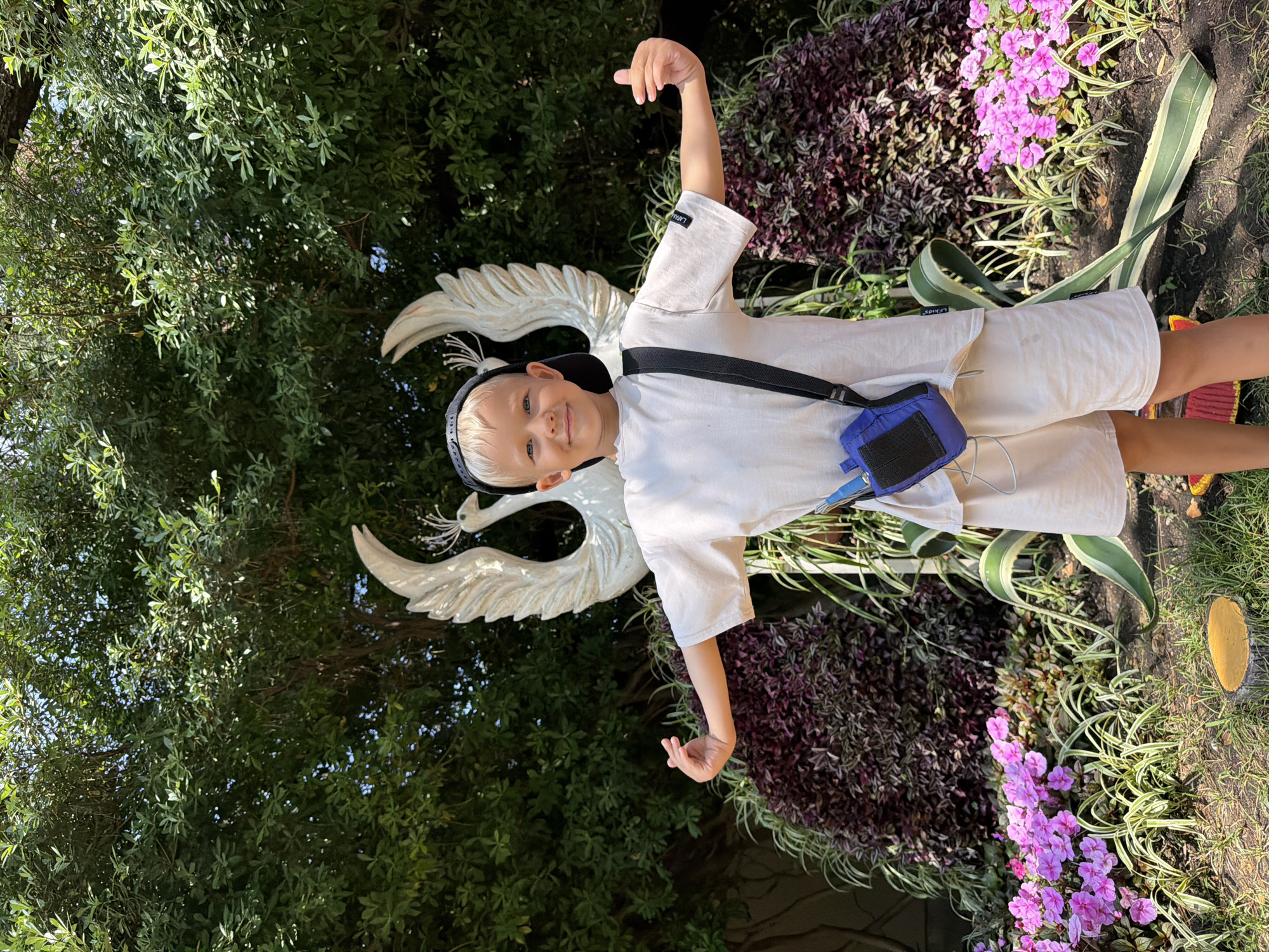 A young boy with blond hair and blue eyes, wearing a white t-shirt, off-white shorts, and a black baseball cap, poses with his arms outstretched in front of a large sculpture of two white birds with spread wings. He has a blue pouch with a black strap hanging across his chest. The background is a lush green garden with various plants, including purple bushes and pink flowers. The lighting suggests it is a sunny day. The location is likely an outdoor public space, possibly a park or garden.