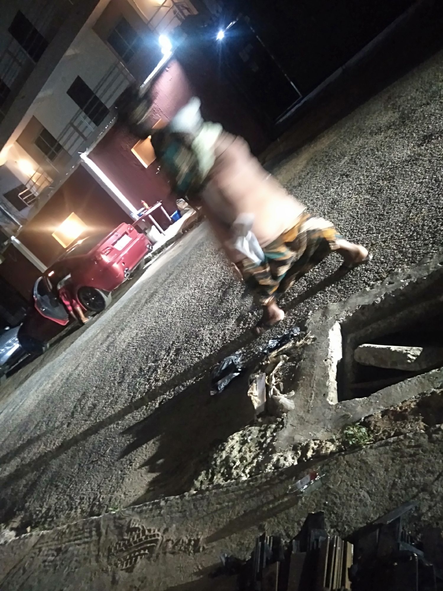 This low-light, blurry photograph captures an outdoor night scene, likely a street or residential area, at a skewed angle. In the center, a heavily motion-blurred person is depicted mid-stride or in a dynamic pose, possibly running or stumbling. The individual appears shirtless, wearing patterned shorts or a wrap-around garment in shades of brown, green, and white, and is barefoot.
To the left, a dark red car, possibly a sedan, is parked with its driver's side door open. Bright reflections from artificial light sources are visible on its surface. In the background, multi-story buildings with illuminated windows and balconies are present, casting light onto the scene.
The ground is a coarse, textured surface, possibly gravel or rough asphalt, and appears damp in places. A visible open drain or gutter runs along the right side, containing some discarded items and debris. In the bottom right corner, dark, rectangular objects are stacked, possibly construction materials. Numerous bright artificial lights illuminate the area, creating significant glare and contributing to the overall blurriness. No discernible text is visible in the image.
