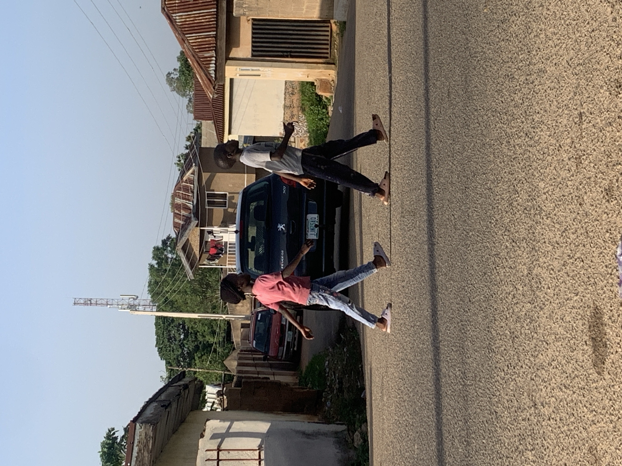 In Jos, Nigeria, two children, a girl and a boy, are walking across a paved road. The girl, on the left, wears a pink t-shirt and blue jeans with white shoes. The boy, on the right, is dressed in a grey t-shirt and dark pants with light-colored slides. They are walking in the same direction, towards the right side of the frame. In the background, a dark-colored car, possibly a Peugeot, is parked on the road. Several residential buildings with corrugated metal roofs and some with concrete walls are visible on either side of the road. A tall telecommunications tower stands in the distance, partially obscured by trees and buildings. The sky is clear and blue, suggesting daylight. The scene appears to be a typical urban or semi-urban setting in Jos.