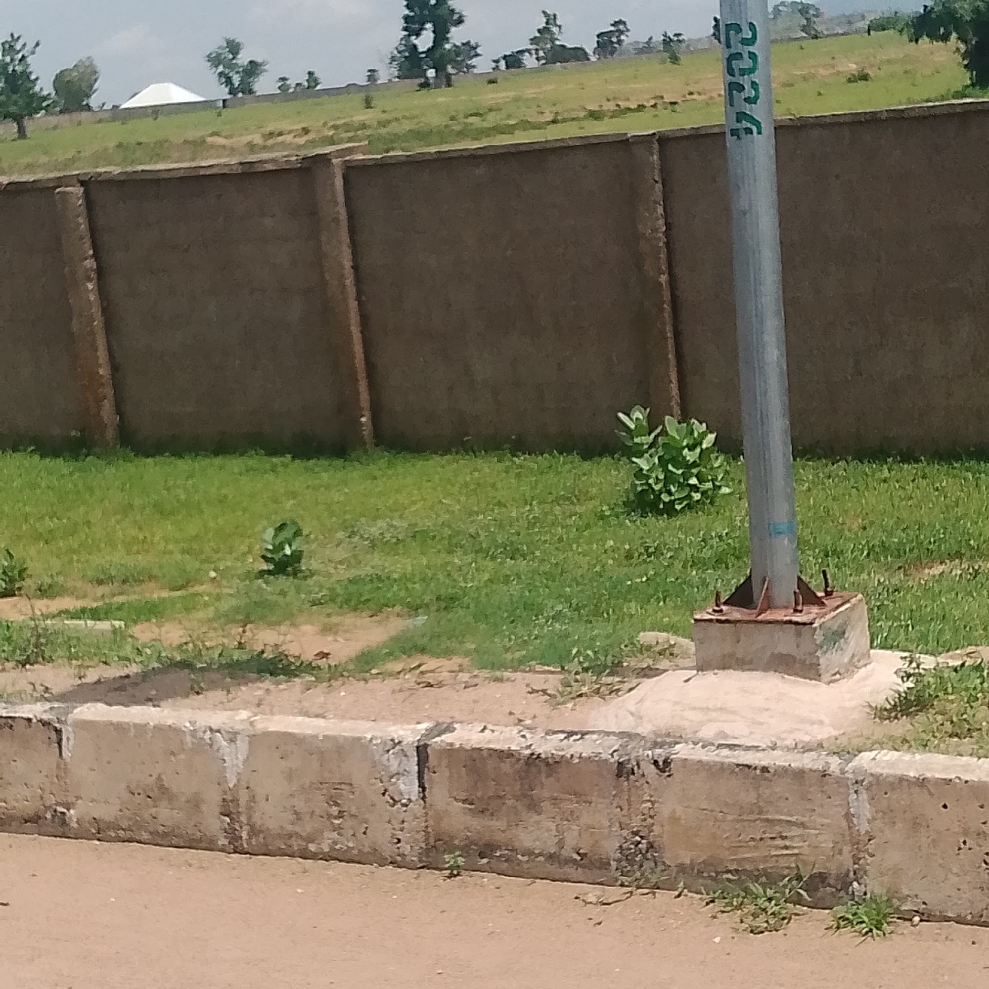 The image depicts a street scene in Jalingo, Nigeria, focusing on a gray metal pole with green markings that appear to be numbers, specifically "2024" vertically oriented. The pole is mounted on a concrete base. In the foreground, a concrete curb lines the edge of a dirt or unpaved road. Behind the curb, a patch of green grass is visible, with some small plants growing within it. Further in the background, a long, low, rough-textured concrete wall stretches across the frame. Beyond the wall, a grassy embankment slopes upwards, dotted with trees. In the far distance, a white, pyramid-shaped structure is visible on top of the hill. The sky is overcast. There are no people or visible signs of activity in the frame.