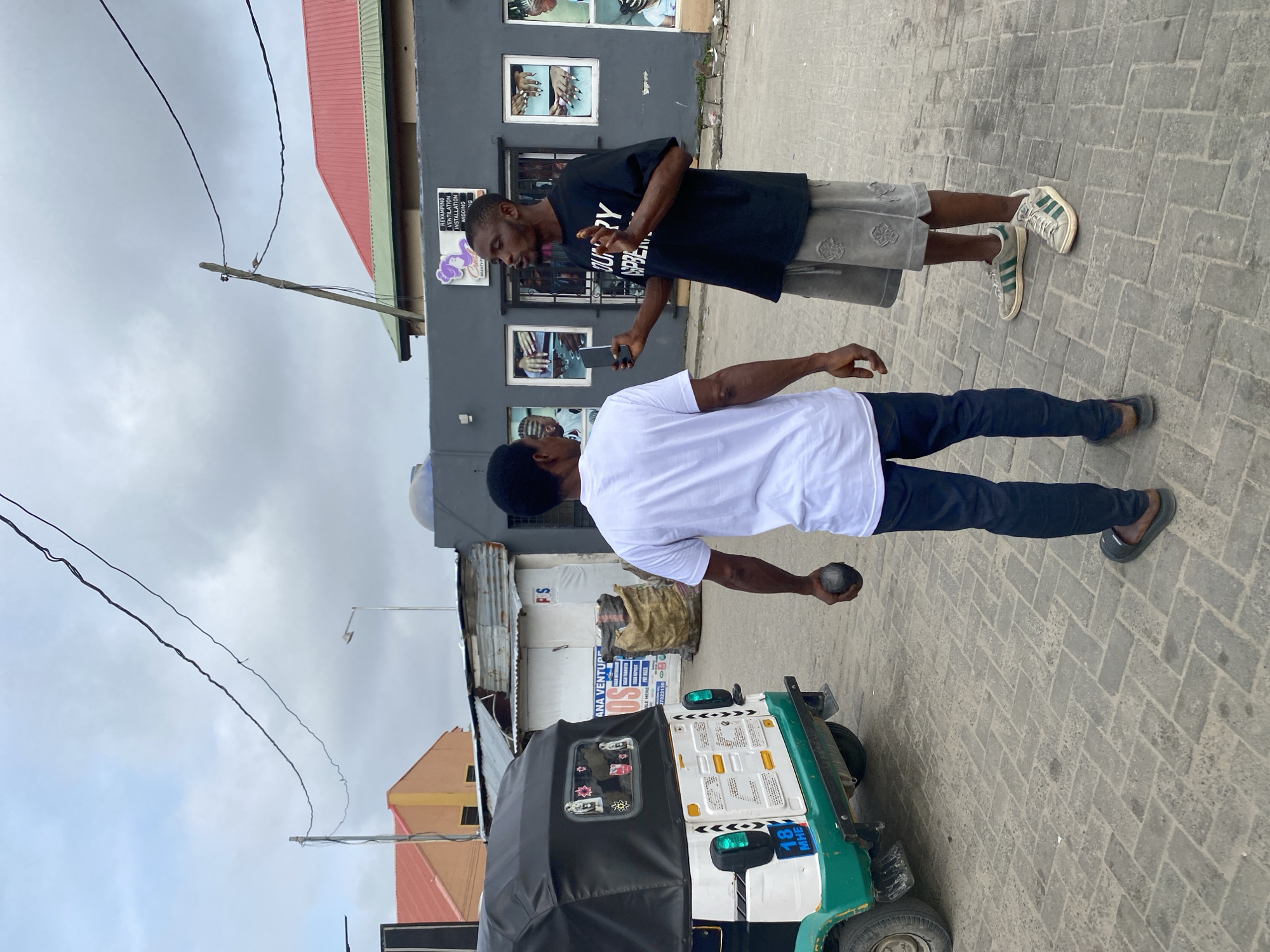 In an outdoor urban setting, two men stand on a paved street under an overcast sky. The man on the left wears a white t-shirt and dark pants, holding a dark, spherical object in his right hand. The man on the right is dressed in a black t-shirt and grey shorts, holding a smartphone in his left hand and gesturing with his right hand as if speaking. A three-wheeled vehicle, partially visible on the left, is white and green. In the background, several buildings are present, one with a red roof and another with a grey facade displaying posters. Overhead, power lines are strung across the sky.