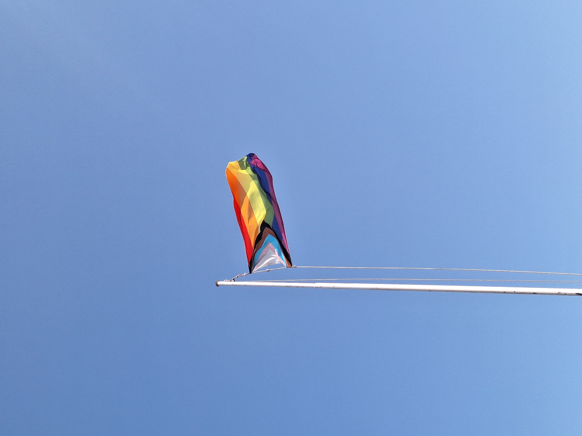 A progressive pride flag and a rainbow pride flag are attached to a white flagpole against a clear blue sky. The flags are flapping in the wind. The flagpole is positioned on the right side of the frame. The image was taken in Arrecife, Spain.