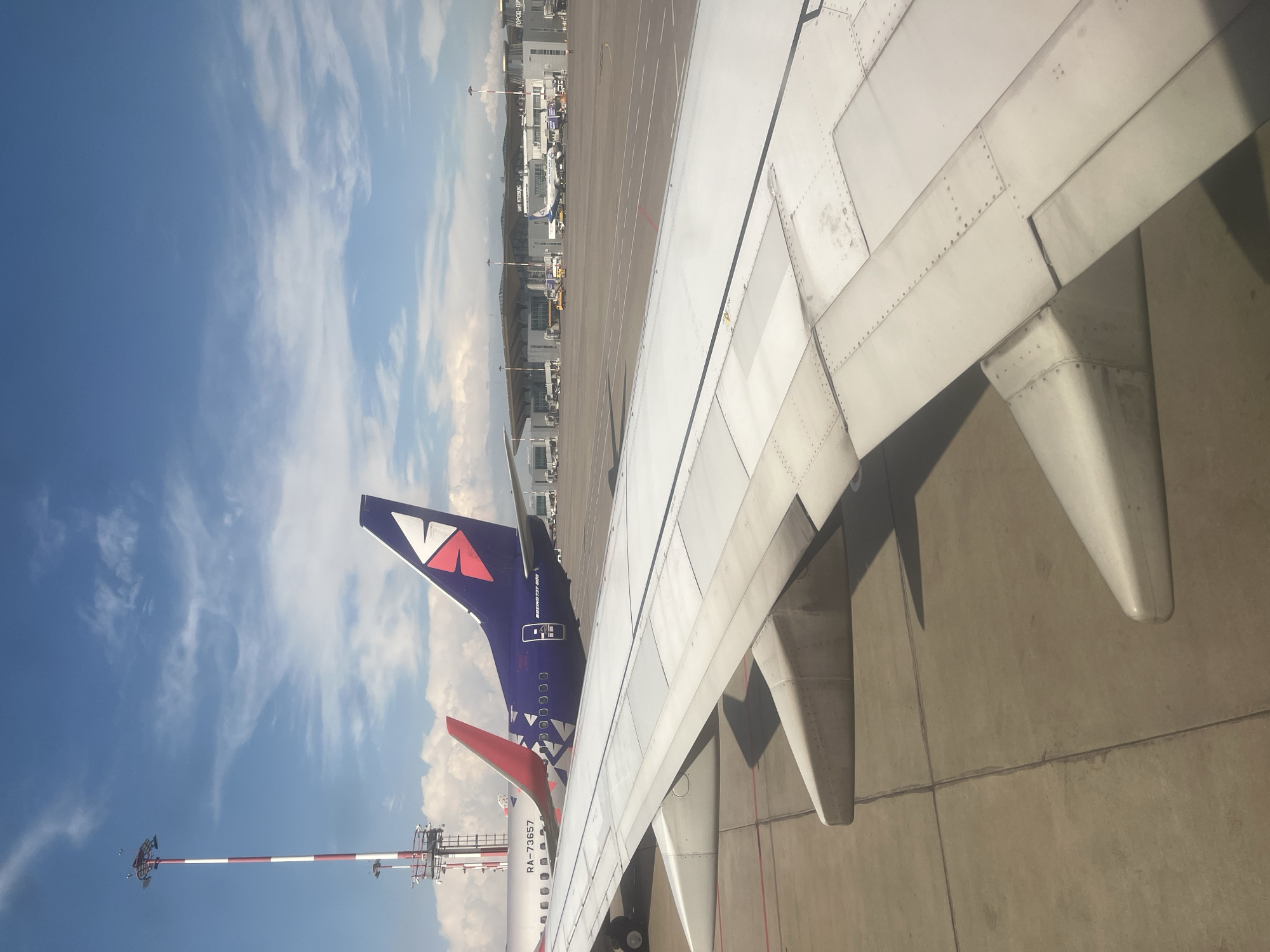 The image captures a view from an airplane window at Pulkovo Airport (LED) in Saint Petersburg, Russia, on a bright, partly cloudy day. The foreground is dominated by the wing of the airplane the viewer is seated in, showing its metallic structure and rivets. In the middle ground, two other airplanes are parked on the tarmac. The tail of a purple airplane with a distinctive white and red logo is prominent, displaying "BOEING 737-800" on its side. To its left, the tail of a white airplane with a red accent and the registration "RA-73557" is visible. In the background, the modern architecture of the airport terminal building is apparent, with its glass facade and roof structure. Several other aircraft and airport service vehicles are scattered across the tarmac. A tall, red and white striped lighting tower stands to the left of the frame, a common sight at airports. The overall scene depicts a typical airport environment, with aircraft ready for departure or arrival under a clear blue sky dotted with scattered white clouds.