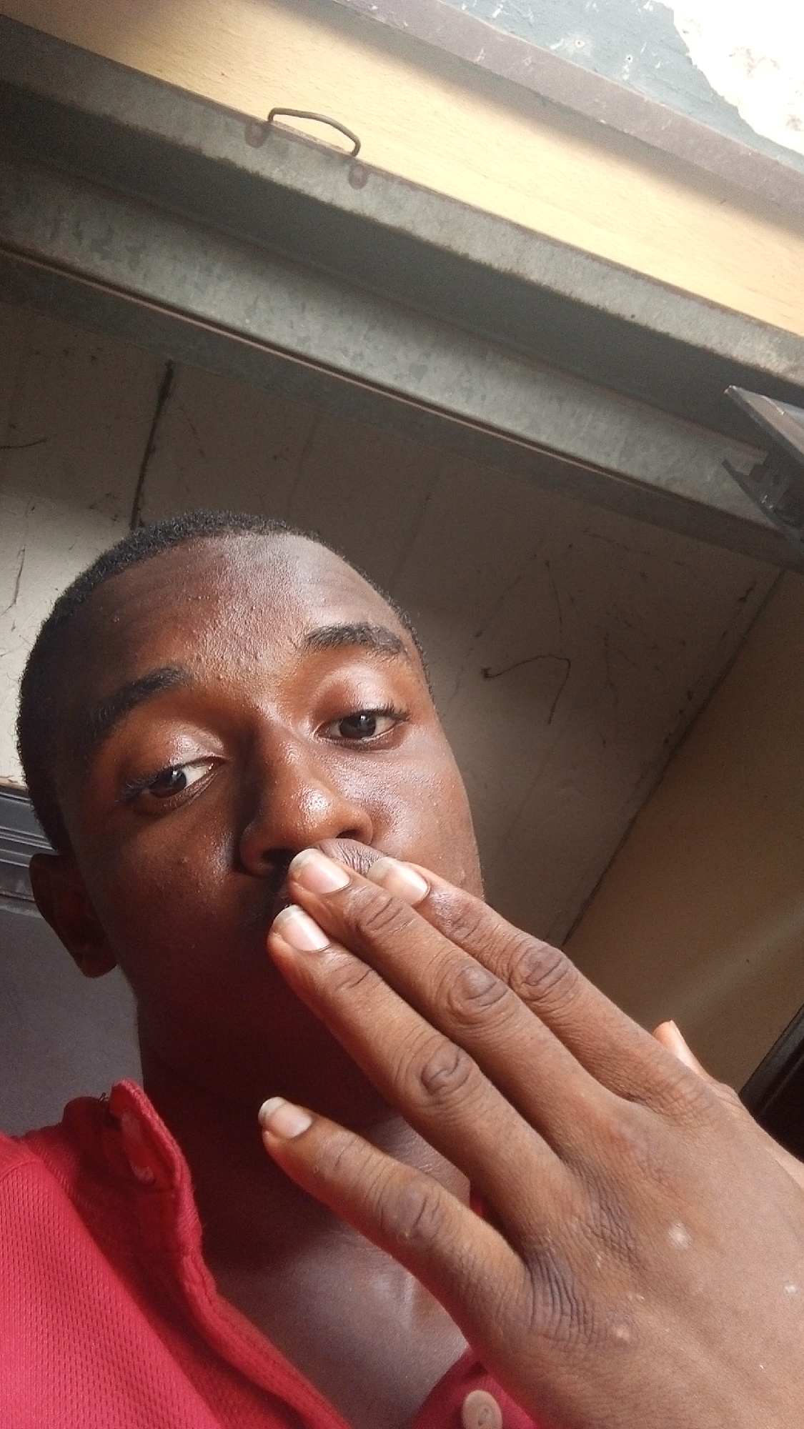 A close-up photograph captures a dark-skinned man in Nkpor, Nigeria, partially obscuring his mouth with his right hand. He is wearing a red collared shirt. The background consists of a metallic and wooden structure, possibly part of a window frame or door. The indoor setting is illuminated by natural light.