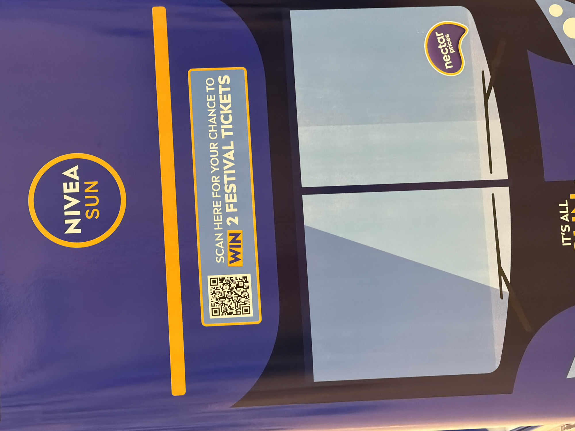 The image shows a portion of a bus advertisement in Pity Me, United Kingdom. The advertisement features the NIVEA SUN logo prominently on the left side. To the right of the logo, there is a call to action: "SCAN HERE FOR YOUR CHANCE TO WIN 2 FESTIVAL TICKETS," accompanied by a QR code for scanning. The central section of the advertisement displays an image that resembles the front windows and windshield wipers of a bus. In the upper right corner, there is a logo that reads "nectar prices". The right edge of the advertisement shows a partial text that reads "IT'S ALL". The background color is primarily a deep blue/purple. The advertisement seems to be promoting a contest or giveaway related to festival tickets in conjunction with NIVEA SUN and nectar prices.