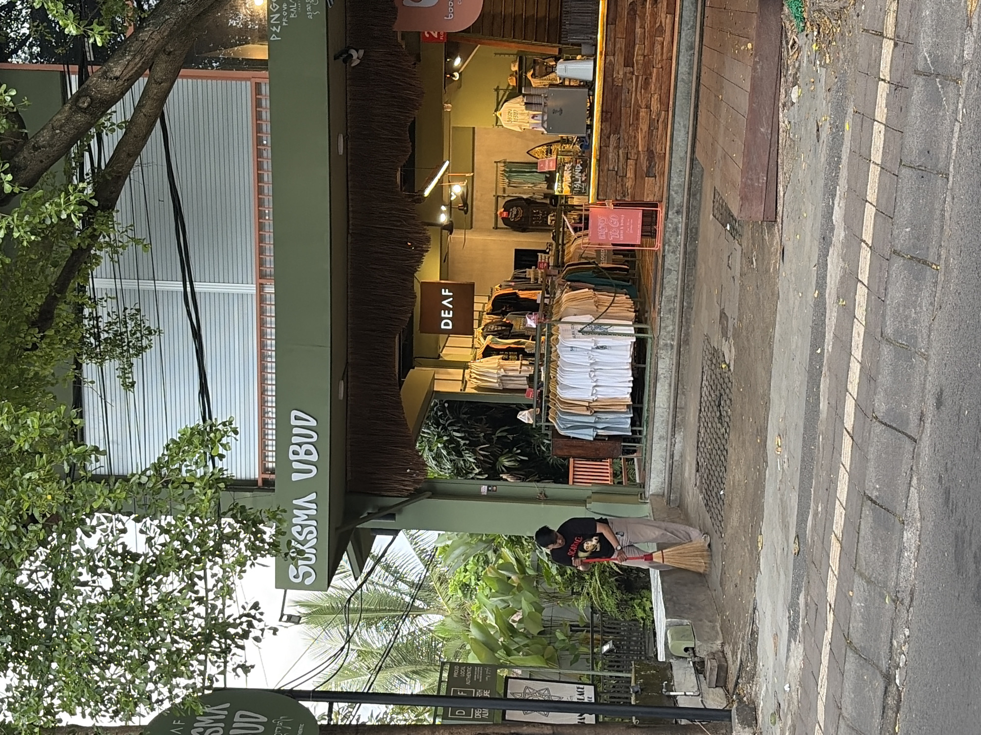 In the city of Ubud, Indonesia, a person is sweeping the sidewalk in front of a store. The store, named "SUKSMA UBUD", has a green facade and a thatched roof overhang. Racks of t-shirts are displayed outside the store. The person sweeping is wearing a black t-shirt with a graphic and tan pants, and is using a broom with a red handle. A tree with green leaves extends over the scene from the left. The sidewalk is made of concrete and pavers.