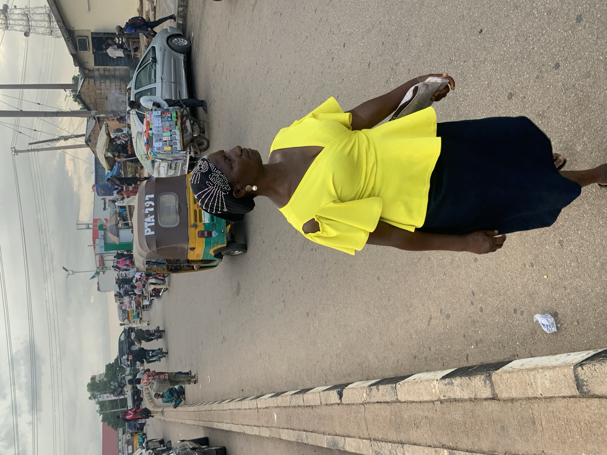 A street scene in Barakin Lerawi, Nigeria, depicts a woman in a bright yellow top and a dark skirt walking on a paved road. She wears a black headscarf adorned with white embellishments and large pearl earrings. To her right, a three-wheeled vehicle, a "Keke Napep," with the number "PTA 191" visible, is on the road. In the background, other vehicles, including motorcycles and cars, are present, along with several people and market stalls lining the street. The environment appears to be a bustling urban area with overhead power lines and some buildings visible. The sky is overcast.