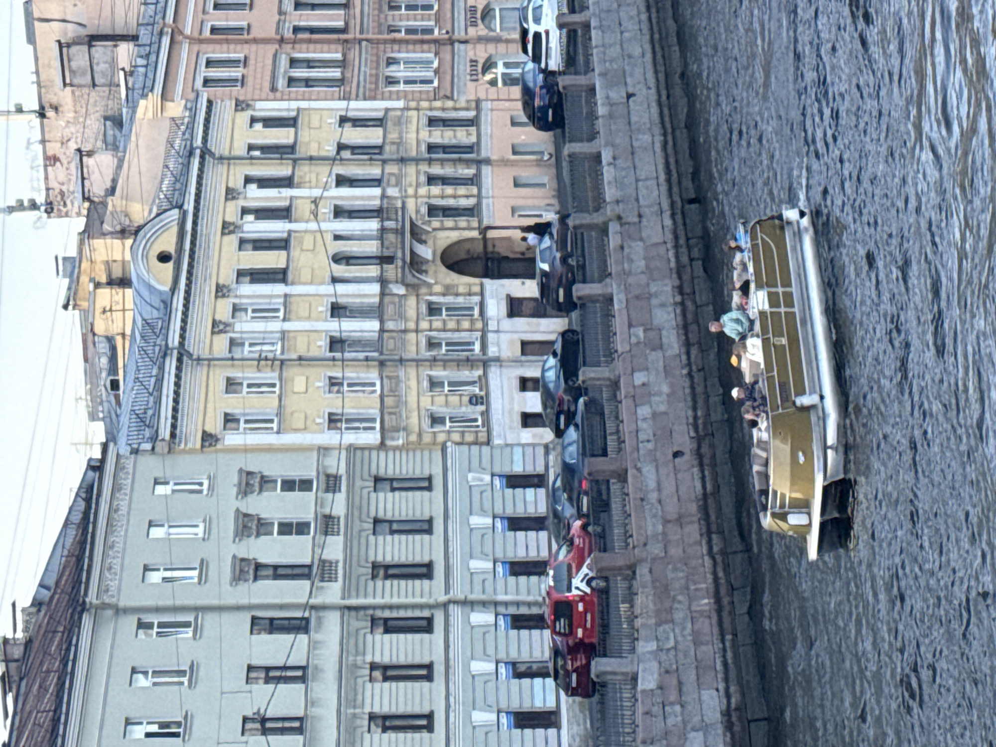 The image shows a canal in Saint Petersburg, Russia, with a wide, flat-bottomed boat carrying several passengers navigating the water. The boat has a golden-colored railing and a white hull, with people seated and standing on deck. On the canal bank, a stone embankment is visible, lined with a decorative fence. Behind the embankment, a row of colorful, multi-story buildings with classical architecture lines the street. Cars are parked along the street in front of the buildings. The buildings are varied in color, including light green, yellow, and pink, and feature numerous windows and architectural details like balconies and ornate moldings. The sky above is overcast. The overall scene depicts a typical urban canal environment in Saint Petersburg, highlighting both the historical architecture and contemporary life.