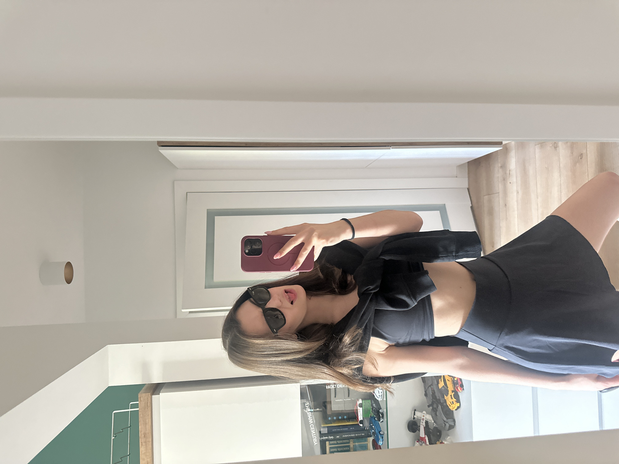 A young female adult is centered in a mirror selfie, captured in an indoor domestic environment in the city of Unknown, Unknown. She has long, dark brown hair, parted on her left, falling over her shoulders. She wears black Wayfarer-style sunglasses and has a playful expression, with her tongue slightly extended and head tilted. Her right hand holds a maroon-cased smartphone with a visible MagSafe ring, taking the photo. Her fingernails are painted pink. A black elastic band is on her right wrist. She is dressed in a black athletic crop top, a black high-waisted flared mini-skirt, and a black sweatshirt tied around her shoulders. Her bare legs are visible.
The background, reflected in the mirror, consists of predominantly white walls, a white ceiling, and light wooden flooring. To her left, a white cabinet with a wooden top and a white wire rack is visible. Books, including one titled "THE BMW CENTURY" (reversed), and several model cars (blue, white, red, yellow, and a grey armored vehicle) are displayed on shelves within the cabinet. A section of a dark green wall is visible above the cabinet. To her right, a plain white wall is visible alongside a white door frame with light blue-grey trim. On the ceiling, a cylindrical white light fixture with a gold interior is mounted. Further back on the right, another white door is partially visible.