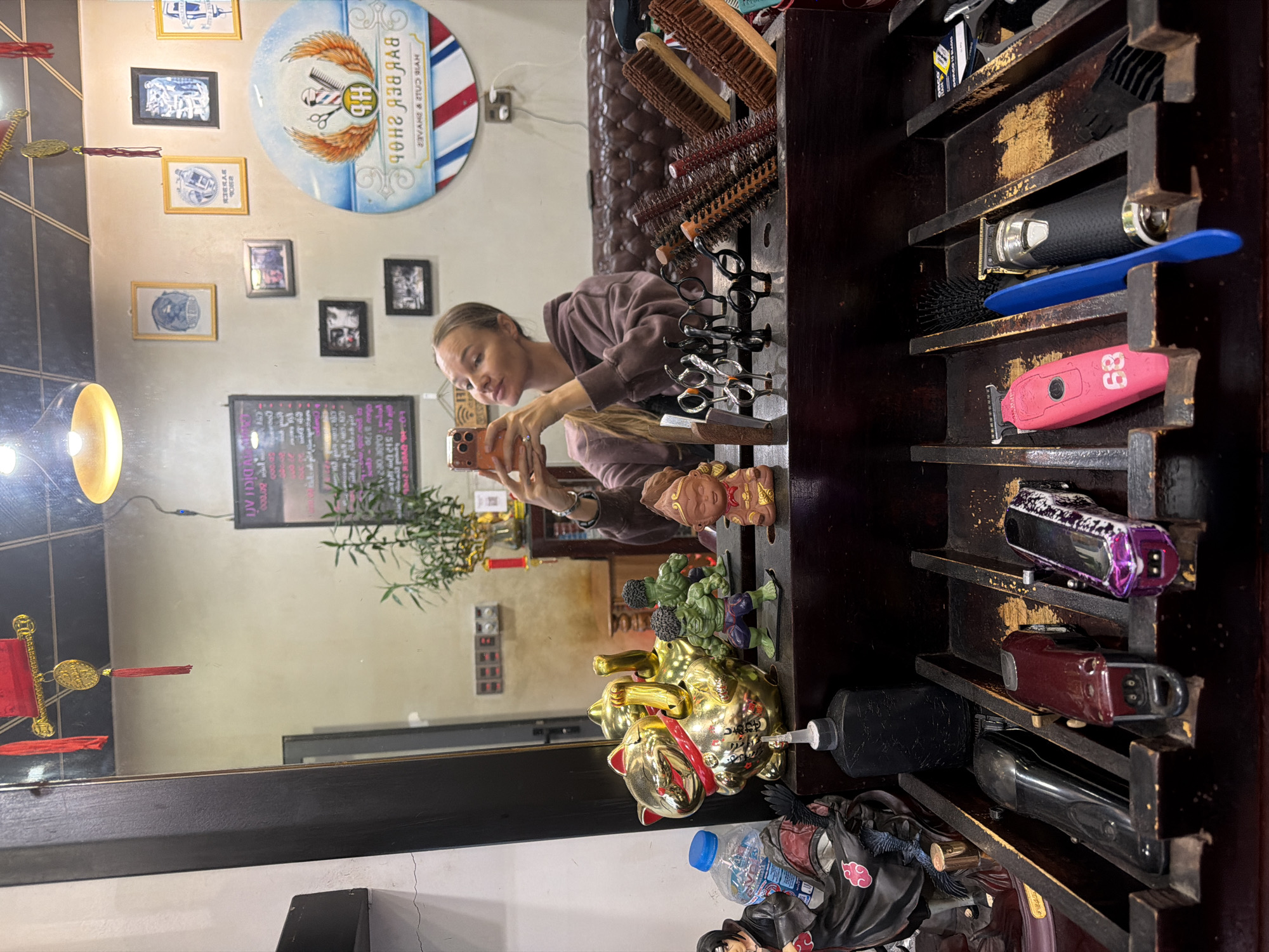 The image shows a woman taking a selfie in a barber shop. She is holding a mobile phone and appears to be looking at its screen. She is wearing a hooded sweatshirt. In the background, there is a mirror reflecting the shop's interior, which includes a barber pole, framed pictures on the wall, and various barber tools such as clippers and brushes. There are also decorative items like a golden cat statue and figurines on a shelf. The shop appears to be located in Xuân Hương - Đà Lạt, Vietnam, as indicated by the Vietnamese text on a sign in the mirror and the style of the decor. The lighting suggests it is daytime.