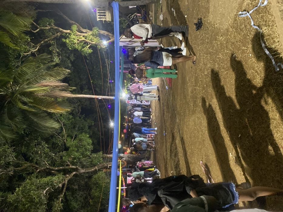 This media file captures an outdoor social gathering at night in Freetown, Sierra Leone. The scene is brightly lit by artificial lights, including spotlights and string lights, which create long, distinct shadows of the attendees on the earthy ground.
Numerous people, mostly young adults, are present. They are seen standing, mingling, and observing, suggesting a relaxed and communal atmosphere. Some individuals in the foreground appear to be sitting or lounging.
A prominent blue fabric banner, possibly with yellow accents, stretches horizontally across the middle of the frame, serving as a decorative element or part of a temporary structure. String lights with small, glowing bulbs are visible, strung along this banner and between the surrounding trees. Large tropical trees, including tall palm trees, flank the gathering space, indicating a natural, open-air setting.
The ground is dry, possibly dirt or sparse grass, with some scattered debris resembling streamers or confetti. In the background, a white drum is faintly visible, hinting at music or entertainment. There is no visible text in the image. The overall impression is that of an evening event or celebration under the open sky.