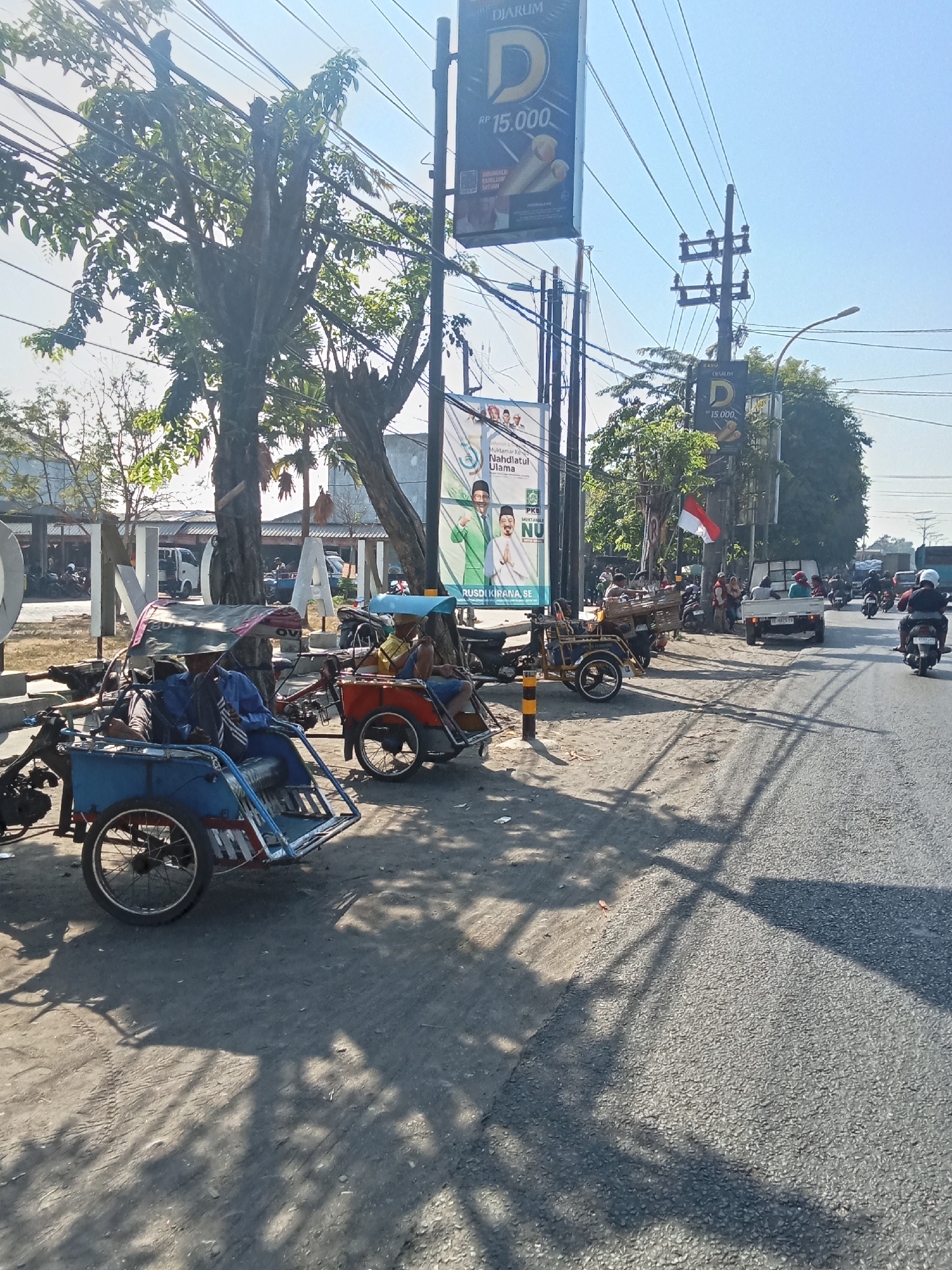 This is a daytime, eye-level, outdoor, medium shot taken on a sunny day on a street in Peterongan, Indonesia. The scene is a roadside with several motorized pedicabs, known as becaks, parked and waiting for passengers. The becaks are colorful, with blue and red predominating. Some have canopies for shade. Several people are seated in the becaks, appearing to be drivers or waiting passengers. A white truck is parked on the side of the road, and other vehicles, including motorcycles, are visible in the background, suggesting a busy street. A tall utility pole with electrical wires runs along the side of the road. Billboards advertising "DJARUM" are also visible. The ground is a mix of dirt and asphalt. Trees with green foliage line the background. The overall impression is a snapshot of local transportation and street life in Indonesia.