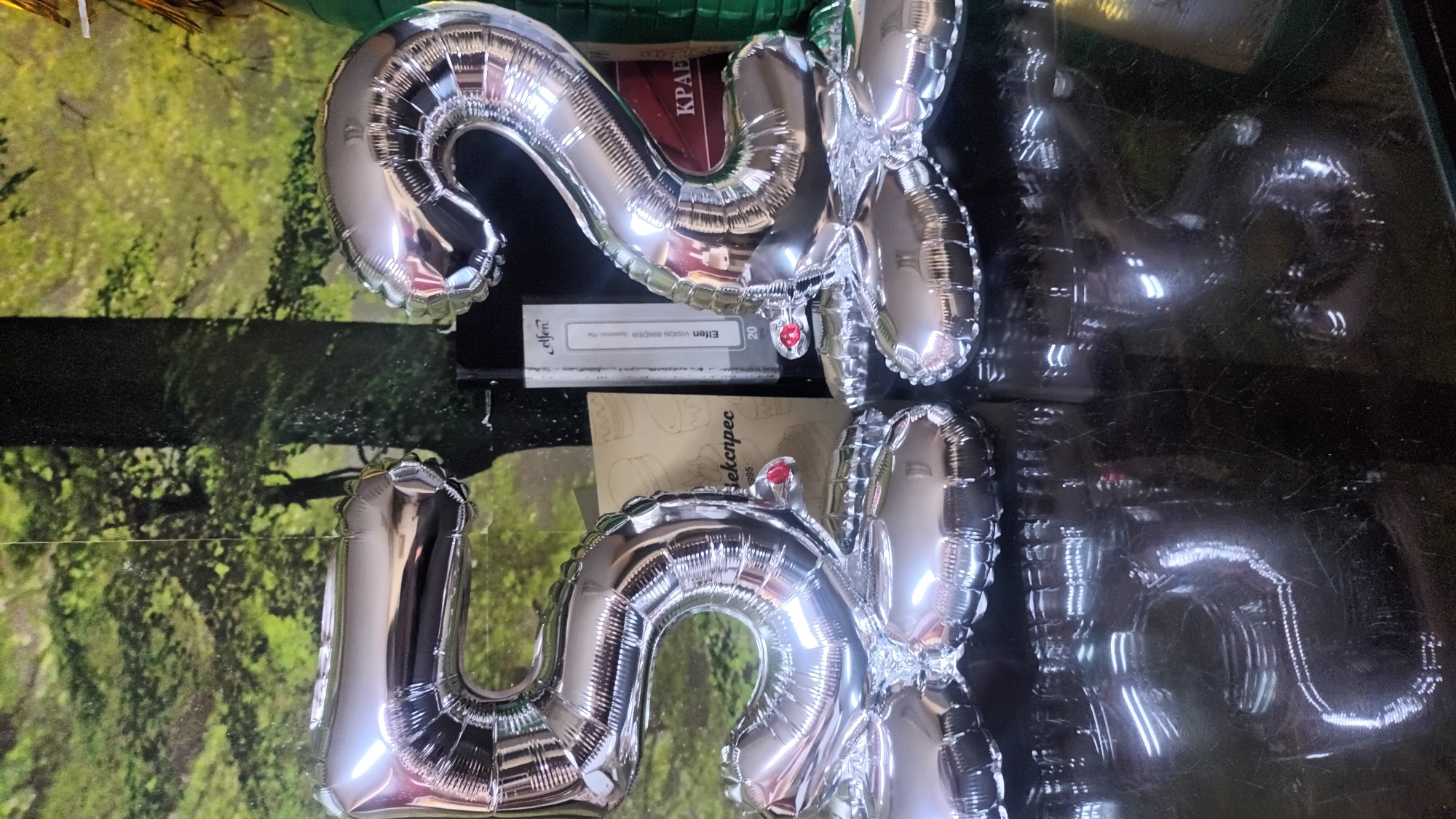 Two silver foil balloons, shaped as the numbers "5" and "2", are positioned in the foreground. The "5" balloon is on the left, and the "2" balloon is on the right. They are resting on a reflective black surface, possibly a table, which shows glare from overhead lighting. Behind the balloons, a backdrop depicts a dense forest scene with green foliage and tree trunks. To the right of the "5" balloon and behind it, there are some objects including a light brown binder or folder with an illustration of cakes and the text "bекnpec" and "1995", and a thin black rectangular object with white text that reads "Elfen VISION BINDER". Further to the right, a red object with white Cyrillic text, possibly "КРАЕВ", is partially visible. The lighting is bright, creating reflections on the balloons and the surface they are on. The location is Varna, Bulgaria.