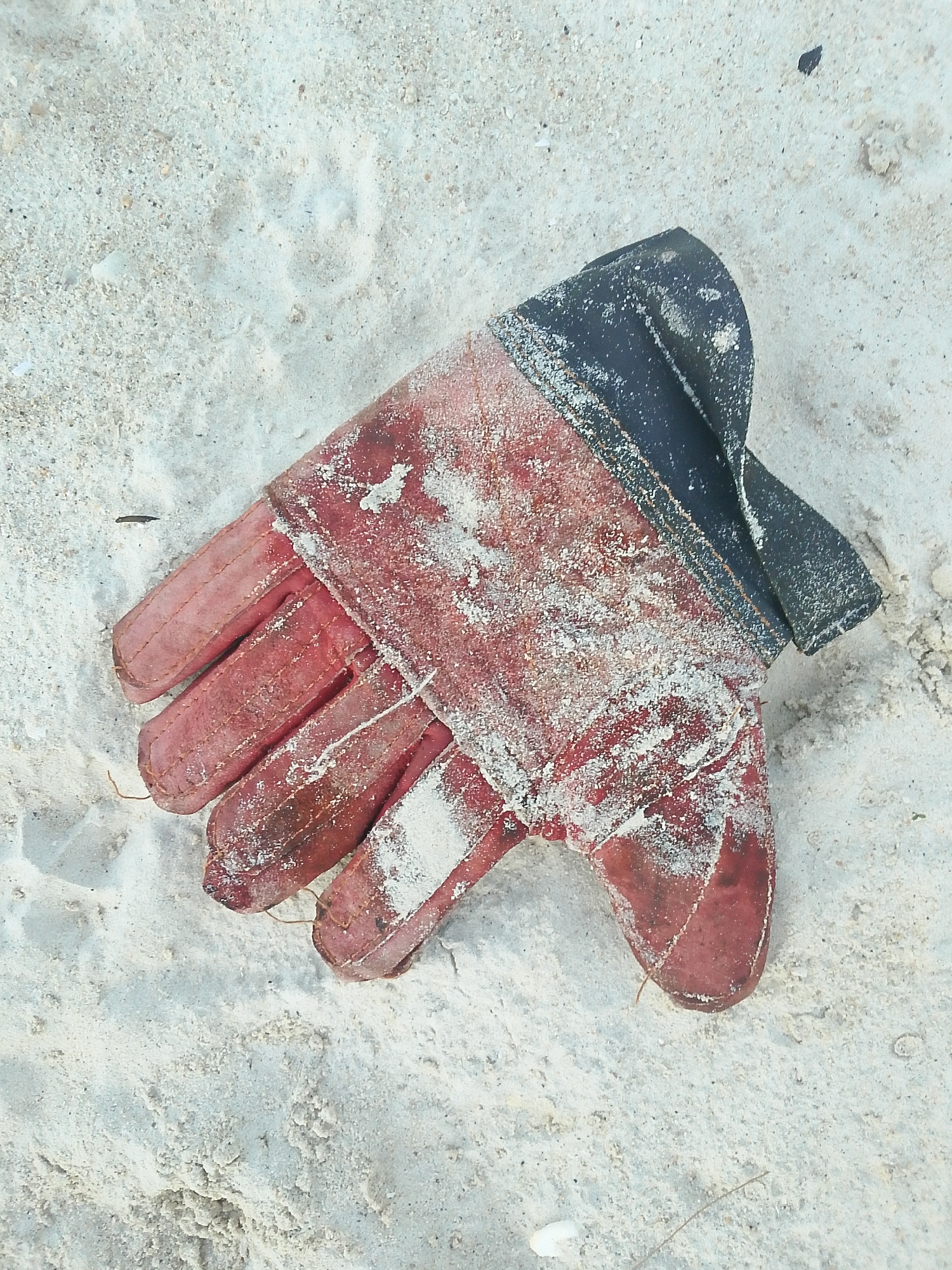 A single red work glove, coated in white sand, lies on a sandy surface. The glove appears to be made of leather and fabric, with a black fabric cuff. The sand is light-colored and granular, with some small debris scattered across its surface. The lighting suggests it is daytime, possibly outdoors. The image does not depict any people, actions, or interactions.