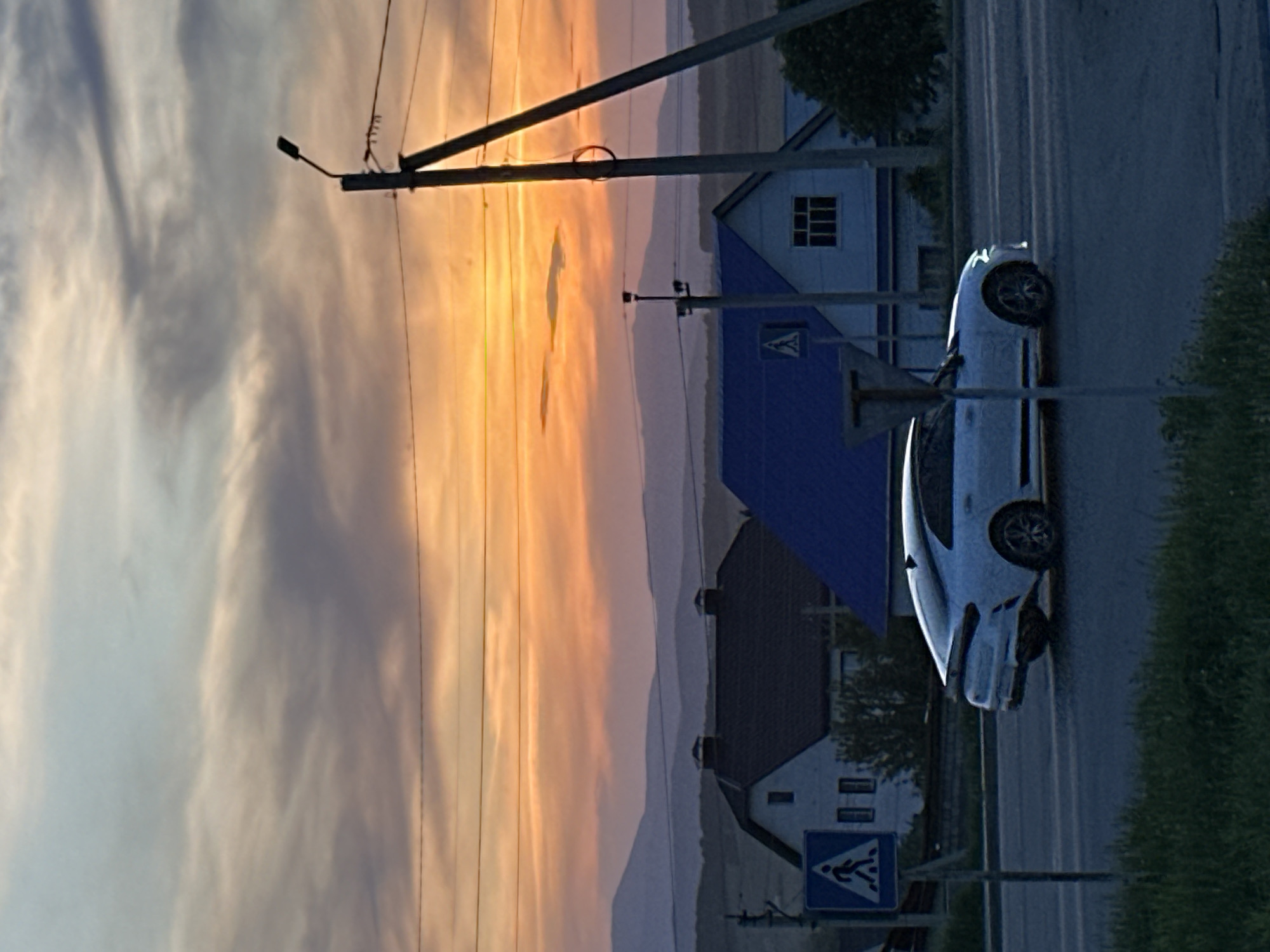 A landscape view shows a car parked on the side of a road. Houses are on the right of the car. The sky is orange to gray with clouds on the left of the car. There are utility poles, wires, and street lights. There is a crosswalk sign close to the houses.