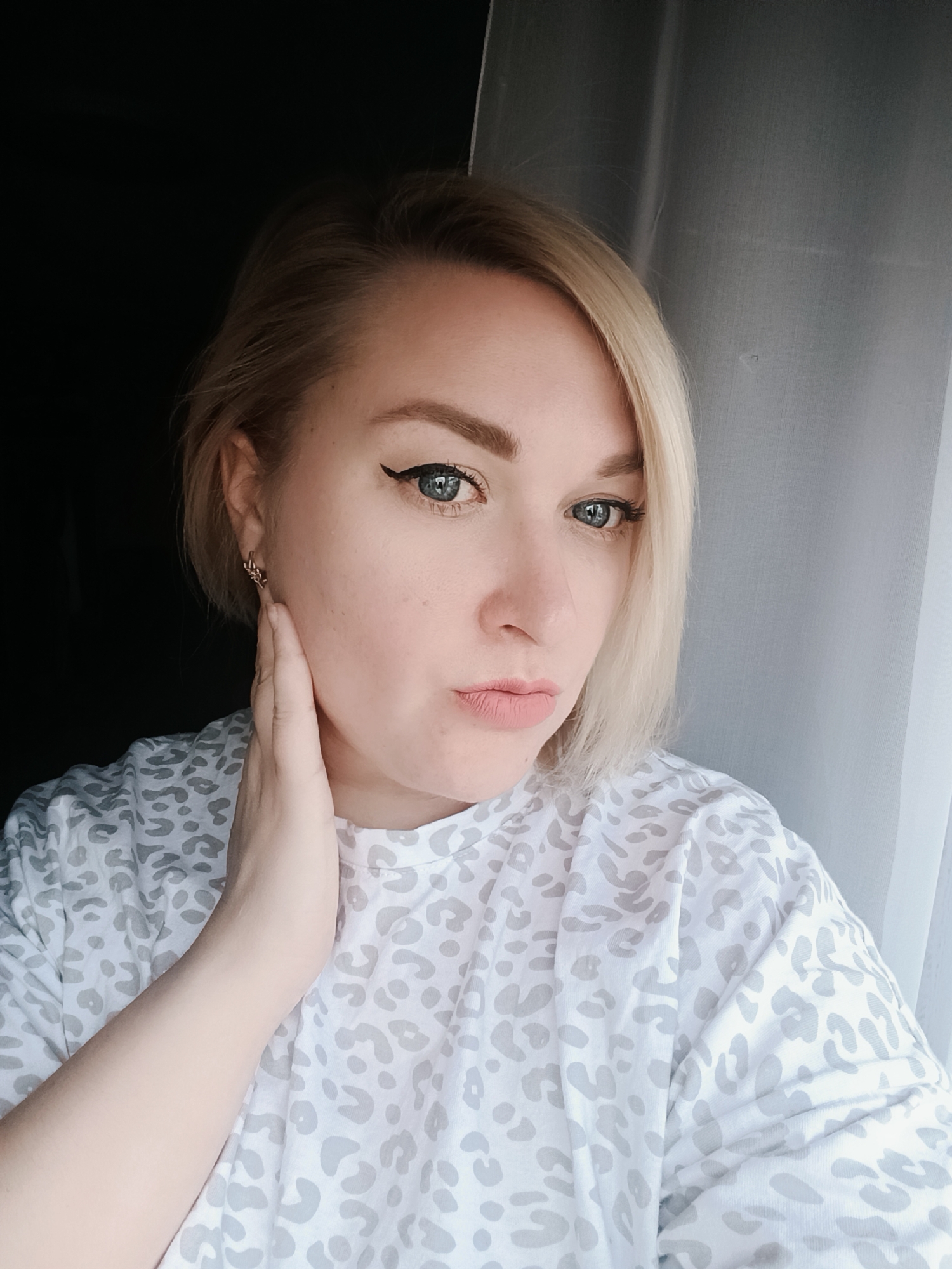 A woman with light blonde, short hair, wearing a white t-shirt with gray leopard print, poses indoors. She is holding her left hand to her neck, facing slightly to the left of the frame. Her makeup includes winged eyeliner. An earring is visible in her left ear. The background includes a dark area to the left and a light-colored curtain to the right. The photo appears to be taken in a room with natural light. There is no specific information available to determine the location in Chaikovsky, Russia.