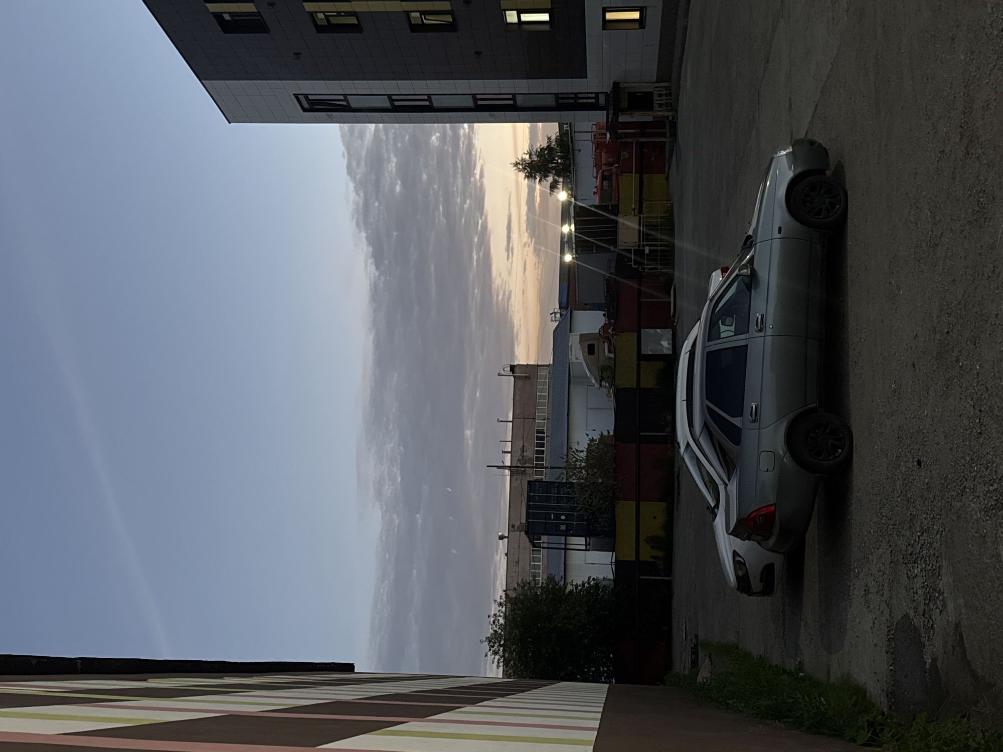 This image captures a twilight scene in an urban industrial area, likely in the city of Perm, Russia, judging by the architectural style and general aesthetic. In the foreground, two cars are parked on a gravel lot. The car closer to the viewer is a silver sedan, facing right, with its rear visible. To its left and slightly behind, a white car is parked, also facing right.
To the left of the parking lot, a building with a distinctive facade of vertical stripes in shades of brown, pink, and light green is partially visible. This building appears to be modern.
In the mid-ground, several industrial structures are present. A dark blue shipping container stands prominently. Behind it, a long, low industrial building with numerous windows stretches across the scene. Further to the right, other industrial elements, including what appear to be trucks or large vehicles, are visible, some painted in bright red and yellow. Several industrial lights are illuminated, casting a glow.
The background is dominated by a wide expanse of sky, displaying the muted colors of dusk. Wispy clouds are scattered across the horizon, and a faint, thin contrail stretches diagonally across the upper portion of the sky. The overall atmosphere is subdued, characteristic of the end of the day in an industrial zone.