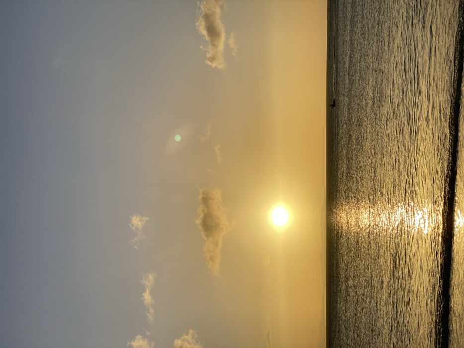 The image displays a coastal sunset scene over the ocean in Juan Griego, Venezuela. The sky transitions from a light blue in the upper portion to a bright golden yellow near the horizon, where a distinct, circular, golden sun is positioned slightly to the left of the image center. Several sparse, white-gold clouds are scattered across the sky, with a notable green lens flare present in the upper right quadrant. The ocean surface, occupying the lower half of the frame, reflects a shimmering vertical path of golden light extending from the sun towards the foreground. A distinct horizon line separates the sky from the water, with faint, distant landmasses visible as low silhouettes. A single, small, dark-colored boat with a solitary figure aboard is observed moving from left to right in the middle ground, to the right of the sun's reflection, leaving a subtle wake. Gentle waves are visible along the immediate foreground shoreline.