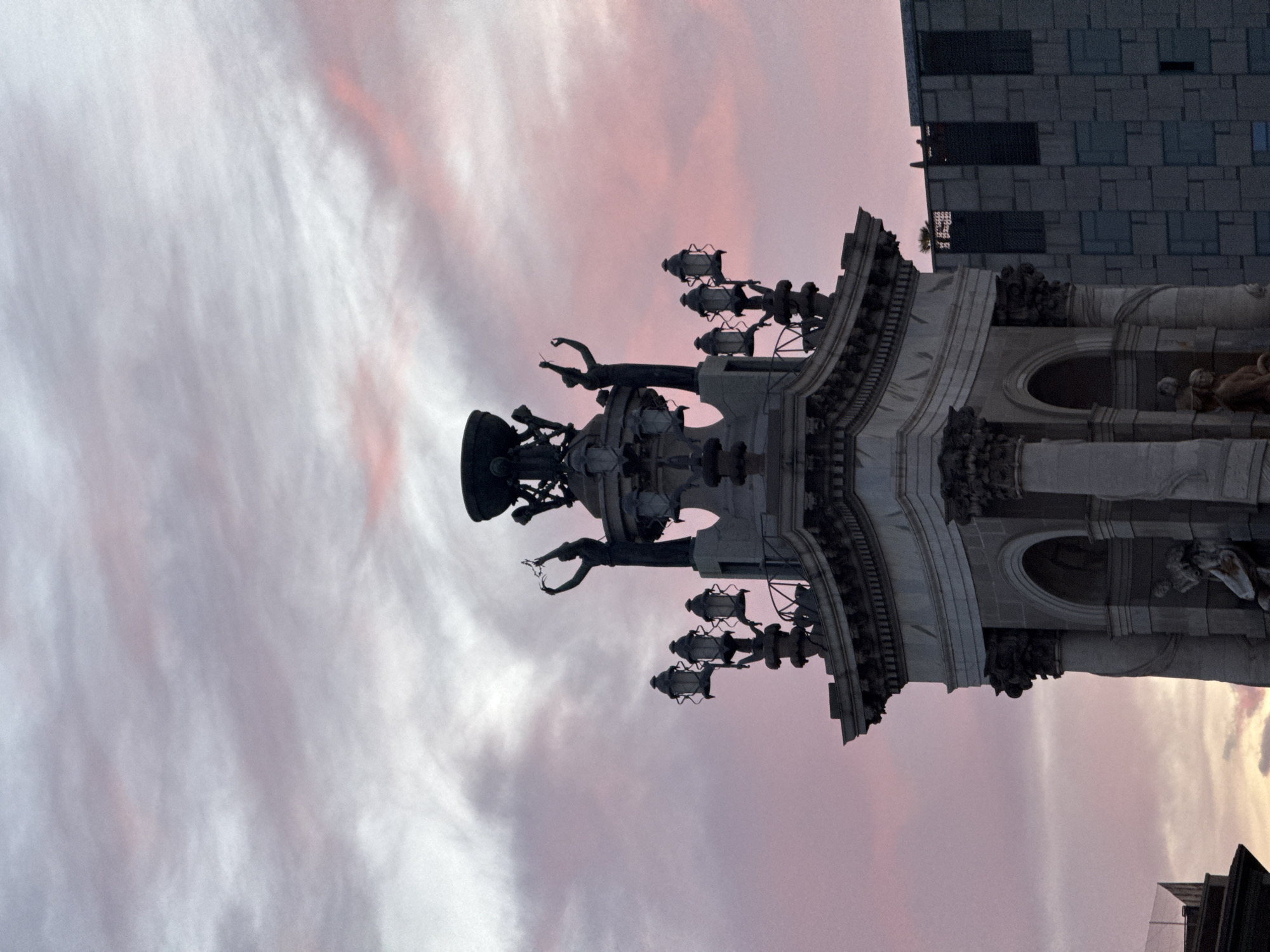 The image shows the Monument a Colom (Columbus Monument) in Barcelona, Spain, set against a cloudy sky with hints of pink. The monument features a column topped with a statue of Christopher Columbus, ornate sculptures, and details. In the background, there is a modern building with a grid-like pattern.