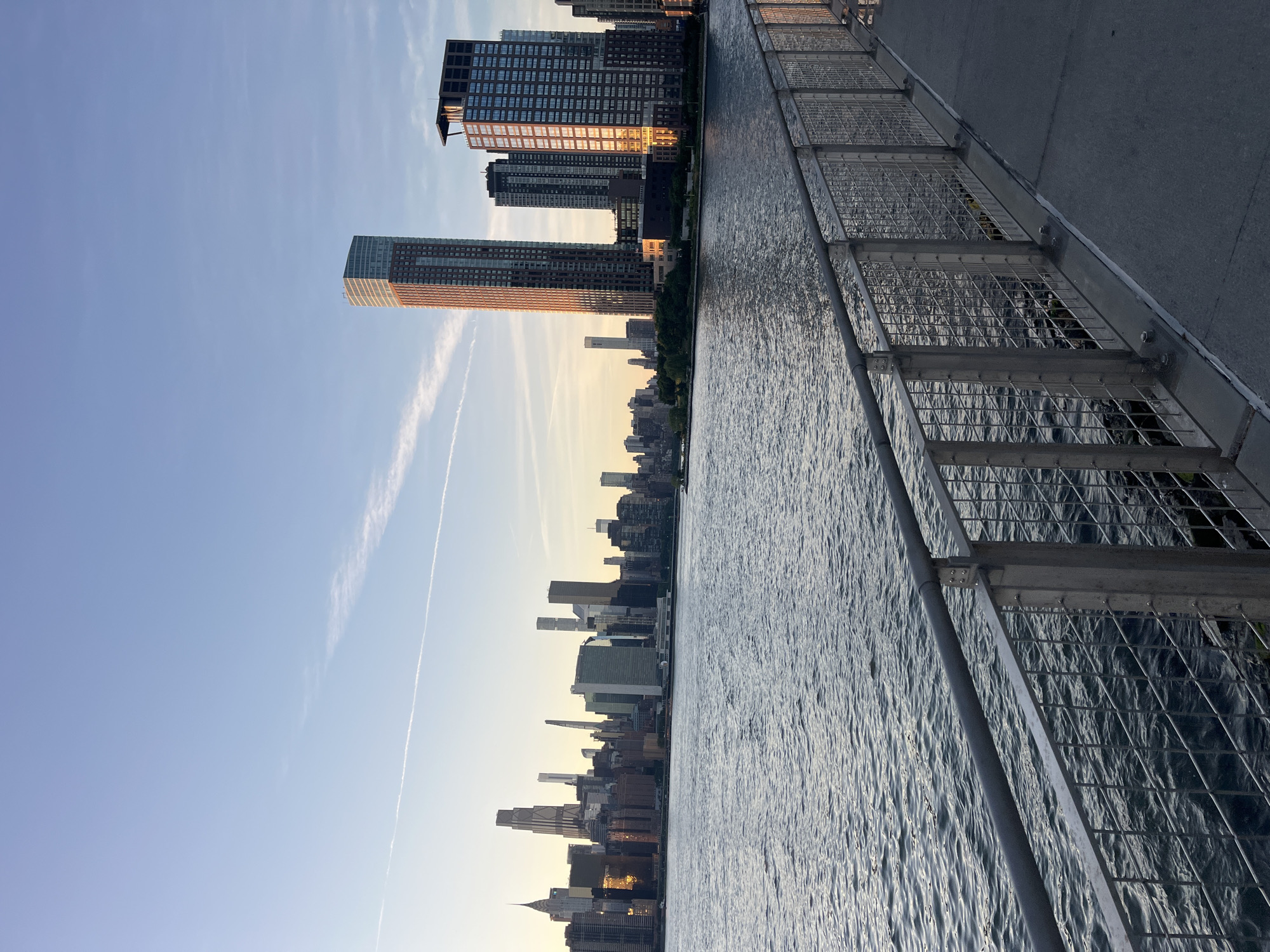 The image shows a view of the Manhattan skyline from across the East River, likely from Brooklyn or Queens, New York City, United States of America. The sun is low in the sky, casting a warm glow on the buildings and water. In the foreground, a paved walkway with a metal railing runs along the waterfront. The water itself is rippled and reflects the light. The skyline features numerous skyscrapers of varying heights and architectural styles. The sky is clear with a few wispy clouds and a contrail from an aircraft. The overall scene is serene and depicts an urban landscape at dusk or dawn.