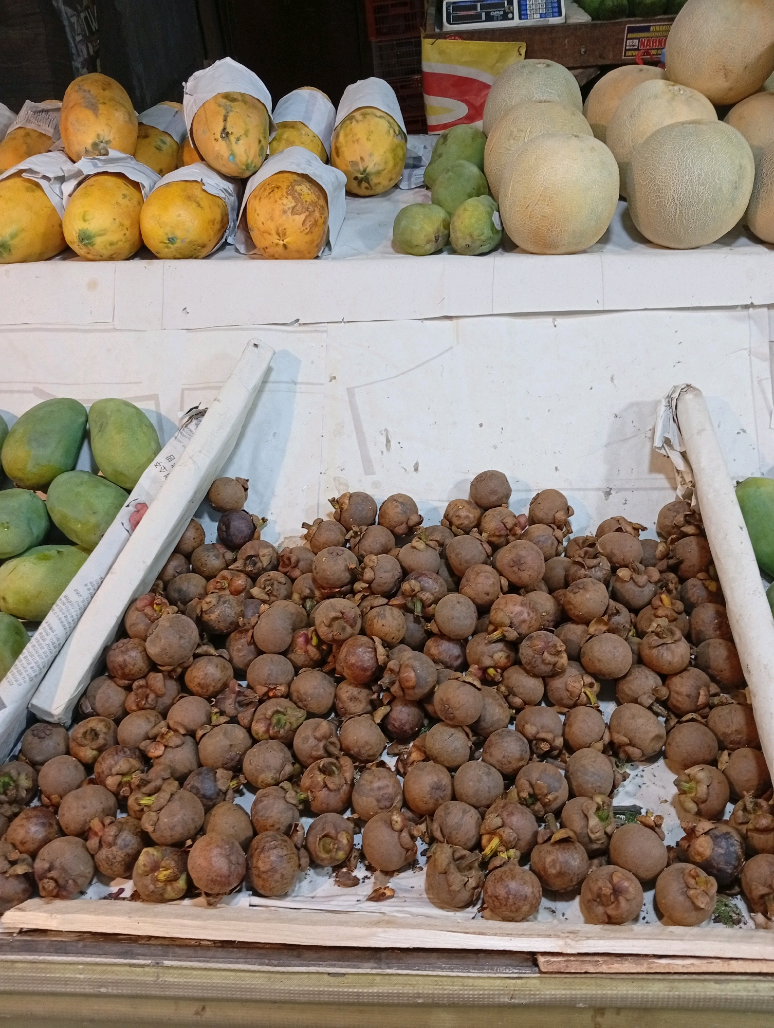A market stall in Kamuning, Indonesia displays an abundance of fresh produce. In the foreground, a large pile of mangosteens, a tropical fruit with a thick, dark purple rind, are neatly arranged. Behind the mangosteens, a collection of green mangoes are visible. Further back, several papayas, some wrapped in paper, are arranged on a white surface. In the top right corner, a variety of melons, including cantaloupes and honeydew melons, are stacked. The lighting suggests an indoor or covered market setting.