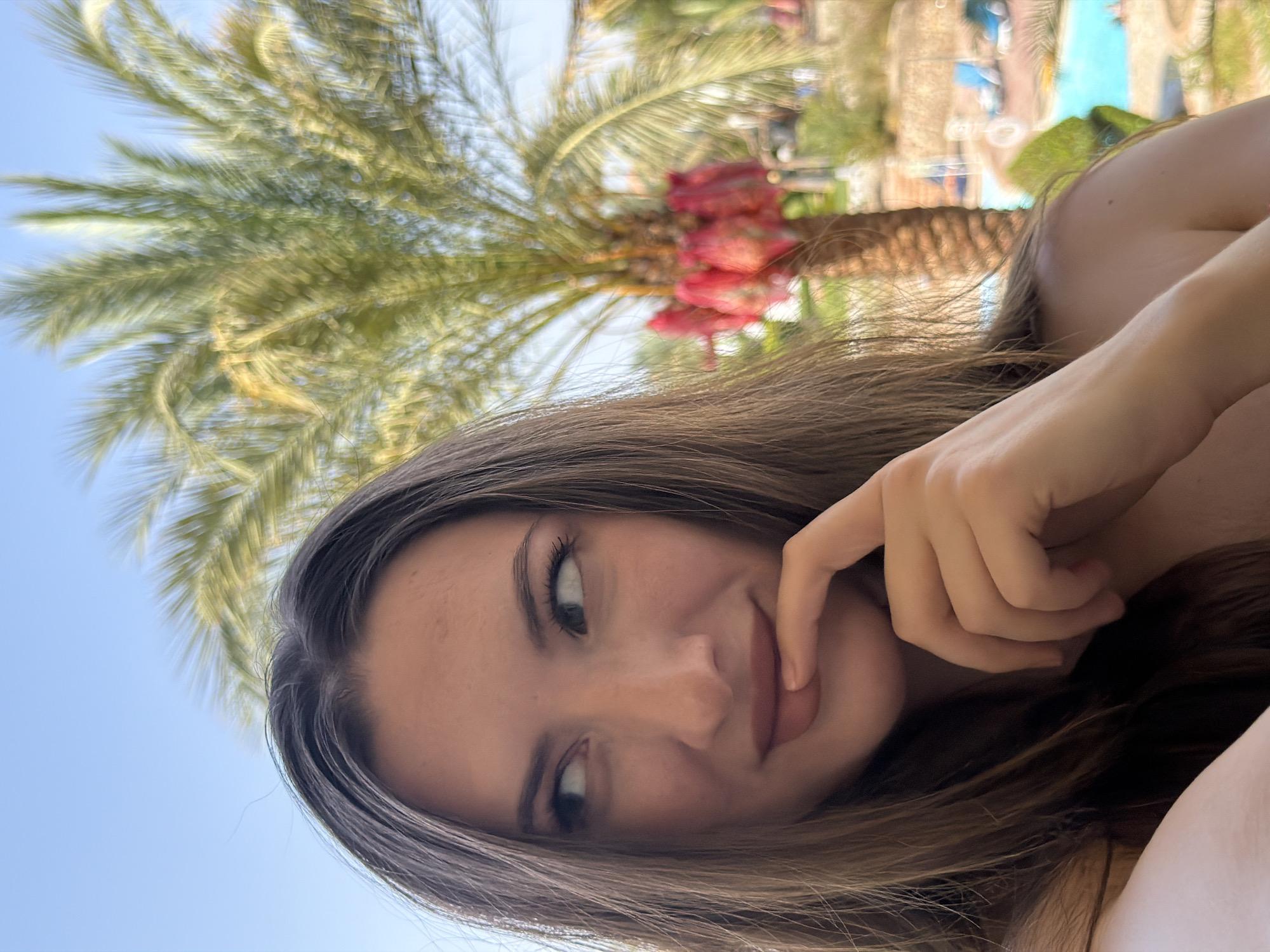 The image captures a close-up of a woman's face and upper body, with a blurred background suggesting an outdoor location. The woman has long, brown hair and is looking off to the left. Her right hand is touching her lips, with her index finger resting on her mouth. She is wearing a nude-colored top.
The background includes a palm tree with red fruits, a bright blue swimming pool with lounge chairs, and other greenery. The overall atmosphere is relaxed and tropical, consistent with a resort in Sharm El Sheikh, Egypt. The lighting indicates it is daytime, likely with bright sunlight.