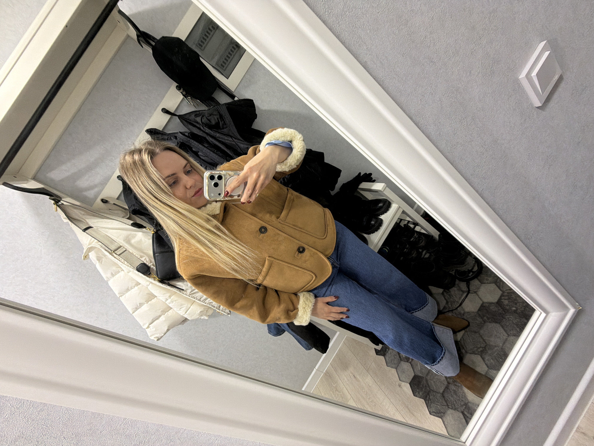 The image displays a blonde woman captured in a mirror selfie within an indoor setting. The specific location within Votkinsk, Russia, is not discernible from the visual information. The environment is consistent with a changing room or a residential entryway.
The woman, appearing to be in her 20s or 30s, is reflected from approximately the chest down, holding a silver-colored smartphone with a transparent case and a triple camera array in her right hand, angled to capture the reflection. She is dressed in a camel-colored, short-length jacket or shearling coat featuring light-colored fleece cuffs and collar, visible buttons, and two front patch pockets. Beneath the jacket, a light blue shirt cuff is visible on her left wrist. She wears blue denim jeans, cuffed at the ankles, and brown ankle boots with pointed toes. Her fingernails are painted dark red.
The large, rectangular mirror has a thick, white beveled frame. In the reflection, a light grey textured wall is visible behind the woman. This wall features a coat rack holding multiple garments, including a white puffer jacket with a black strap and what appears to be a black crossbody bag, alongside several dark-colored jackets or coats. Below the coat rack, a white multi-tiered shoe shelf displays dark footwear. The floor reflected in the mirror transitions from a section of hexagonal grey tiles to light wood-look panels. A white decorative trim runs along the upper part of the wall, and a square white fixture, possibly a light or vent, is mounted on the wall to the far right of the reflection.