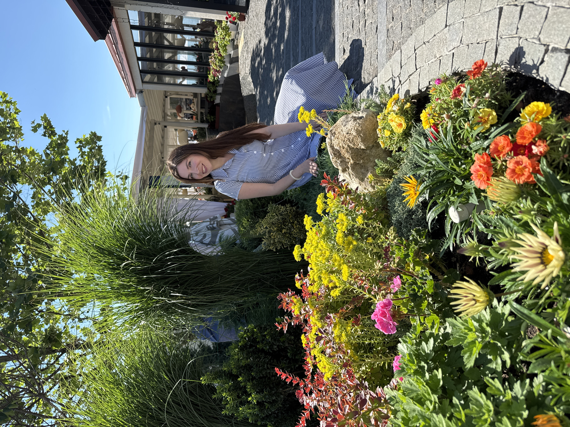 In Odesa, Ukraine, a young woman with long, brown hair sits on a grassy area beside a vibrant flowerbed filled with red, yellow, and pink blossoms. She is wearing a blue and white striped dress and smiles at the camera. Behind her, tall green ornamental grasses frame the scene, and in the distance, a building with large windows and awnings suggests a restaurant or cafe, with tables and chairs visible through the glass. The sun shines brightly in a clear blue sky, casting shadows on the cobblestone path that curves around the flowerbed.
