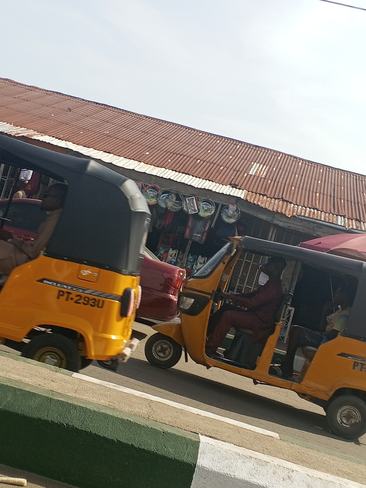 The image shows a street scene in Jalingo, Nigeria, with two yellow auto-rickshaws, also known as "keke napep," driving on a road. The auto-rickshaws are open-sided vehicles with canopies. The first auto-rickshaw, on the left, has a black canopy and is partially visible. A person wearing glasses and a light brown garment is seated in the driver's position. The rear of this auto-rickshaw displays the license plate "PT-293U". Behind it, a red car is also visible.
The second auto-rickshaw, on the right, also has a black canopy and a red umbrella partially obscuring the top. A person wearing a dark red outfit is in the driver's seat, steering the vehicle. Another person, partially visible, is seated behind the driver. This auto-rickshaw also has a license plate which is partially visible.
In the background, a row of market stalls with corrugated metal roofs is visible. Various items, possibly merchandise, are displayed under the awnings of the stalls. The sky is bright and clear. The road in the foreground has a green and white curb. The overall impression is of a busy urban street environment in Nigeria.