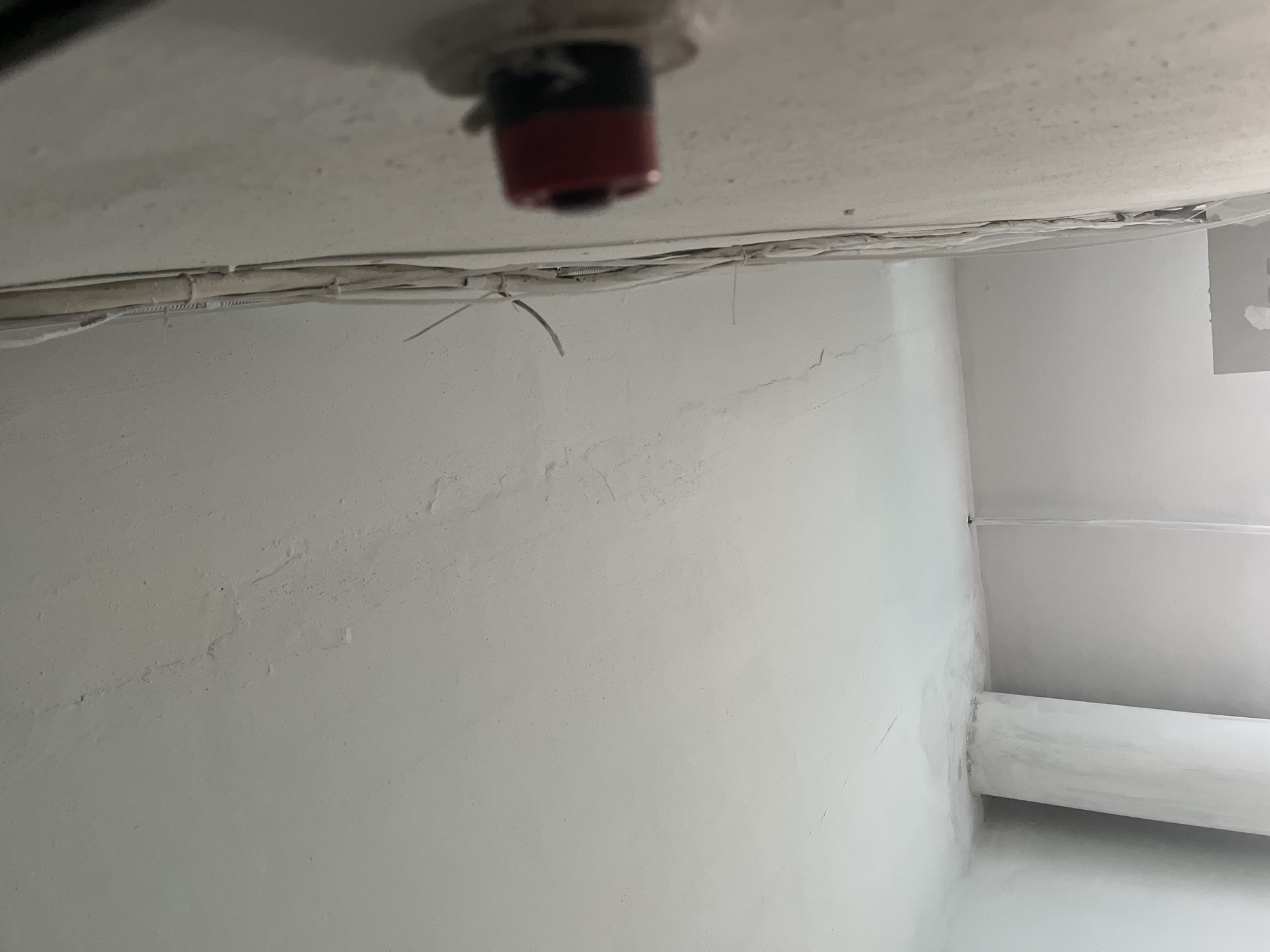 An indoor image shows a close-up of a textured white ceiling with some hairline cracks visible. To the right, a white wall features a bundle of electrical wires running vertically, secured with ties. A red and black electrical connector or component is attached to the wall, partially obscuring the wires. In the lower left portion of the image, a white, cylindrical pillar or column extends upwards and appears to be connected to the ceiling. The background suggests a room under construction or renovation, with unfinished surfaces. The lighting is diffuse, casting soft shadows.