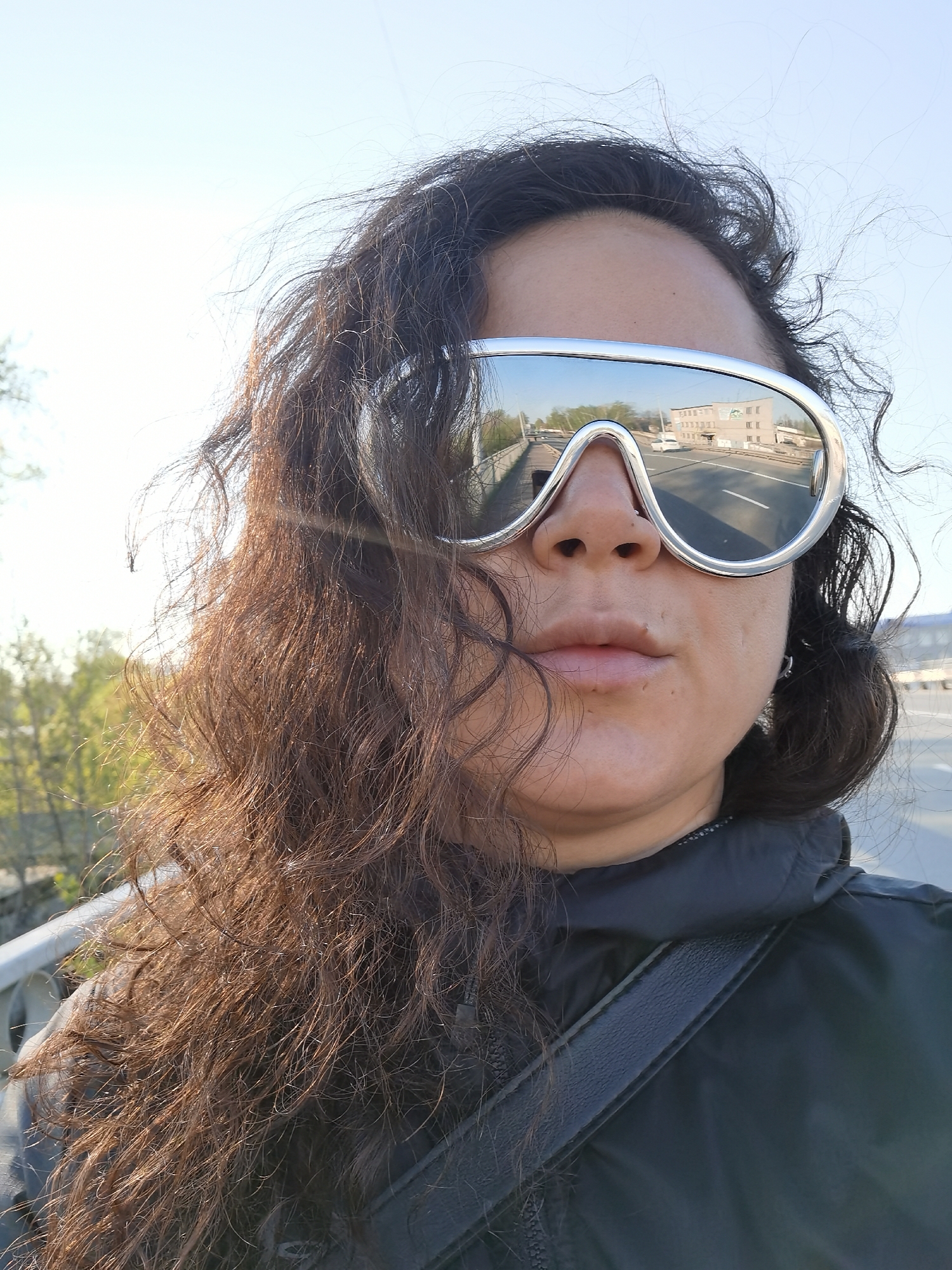 A selfie of a person with curly, brown hair and silver, mirrored sunglasses, taken outdoors in Chaikovsky, Russia. The reflection in the sunglasses shows a road, a white car, and a building. The person is wearing a dark jacket with a strap across their chest. The background suggests they are on a bridge or elevated walkway, with trees and a sky visible in the distance. The photo is taken in daylight, appearing to be a clear day.