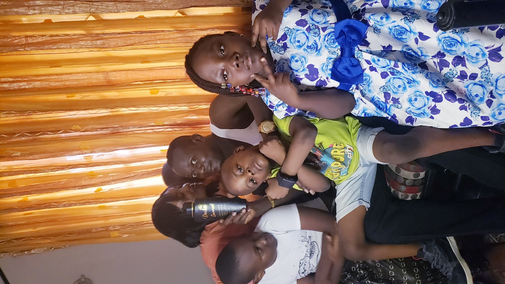 A group of six people are indoors in what appears to be a home in Sabon Tasha, Nigeria. An adult male stands near the center of the group. He wears a white tank top and a gold watch. To his left, a woman holds a black bottle labeled "Duba Cameroon". To his right, a child in a green shirt wearing a black smartwatch looks at the camera. A child wearing a white shirt is seated. A child wearing a white and blue dress raises a hand in a peace sign. Orange curtains are visible behind them.