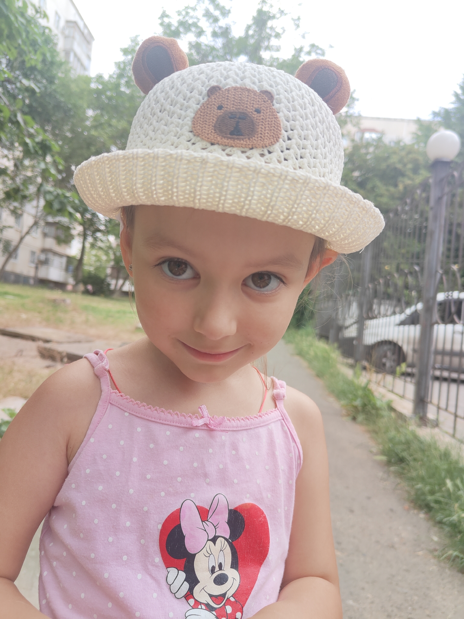 A young girl is captured in a close-up, outdoor portrait. She is wearing a white woven hat adorned with brown bear ears and a brown bear face. The hat has a wide brim. She is also wearing a pink tank top with white polka dots and a graphic of Minnie Mouse within a heart on the front.
The girl is looking directly at the camera with a slight smile. Her eyes are brown, and her hair appears to be light brown. She has a small silver earring in her visible ear. The background is slightly blurred, suggesting a shallow depth of field.
The setting appears to be a sidewalk or path, with a metal fence and green grass visible to the right. In the distance, there are trees, a building, and a parked white car. The lighting suggests it is daytime, possibly overcast due to the soft light. The overall mood is casual and sweet, focusing on the child's playful attire and expression. The location context provided is Chornomorsk, Ukraine. No visible text is present in the image.