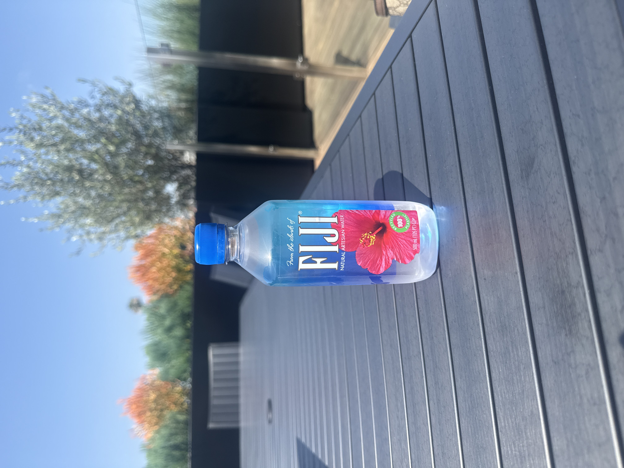 A bottle of FIJI water rests on a gray outdoor table with slatted surface. The background shows a view of trees and a clear blue sky. The label of the bottle includes text that reads "From the islands of FIJI" and "NATURAL ARTESIAN WATER," along with a red flower graphic. The text also shows that the bottle is made of "100% Recycled Plastic" and "500 ml (1.05 PT)." Based on the background vegetation and clear sky, the scene likely takes place in a residential or recreational outdoor area, possibly in Los Angeles, United States of America, but there is no concrete information to confirm this.