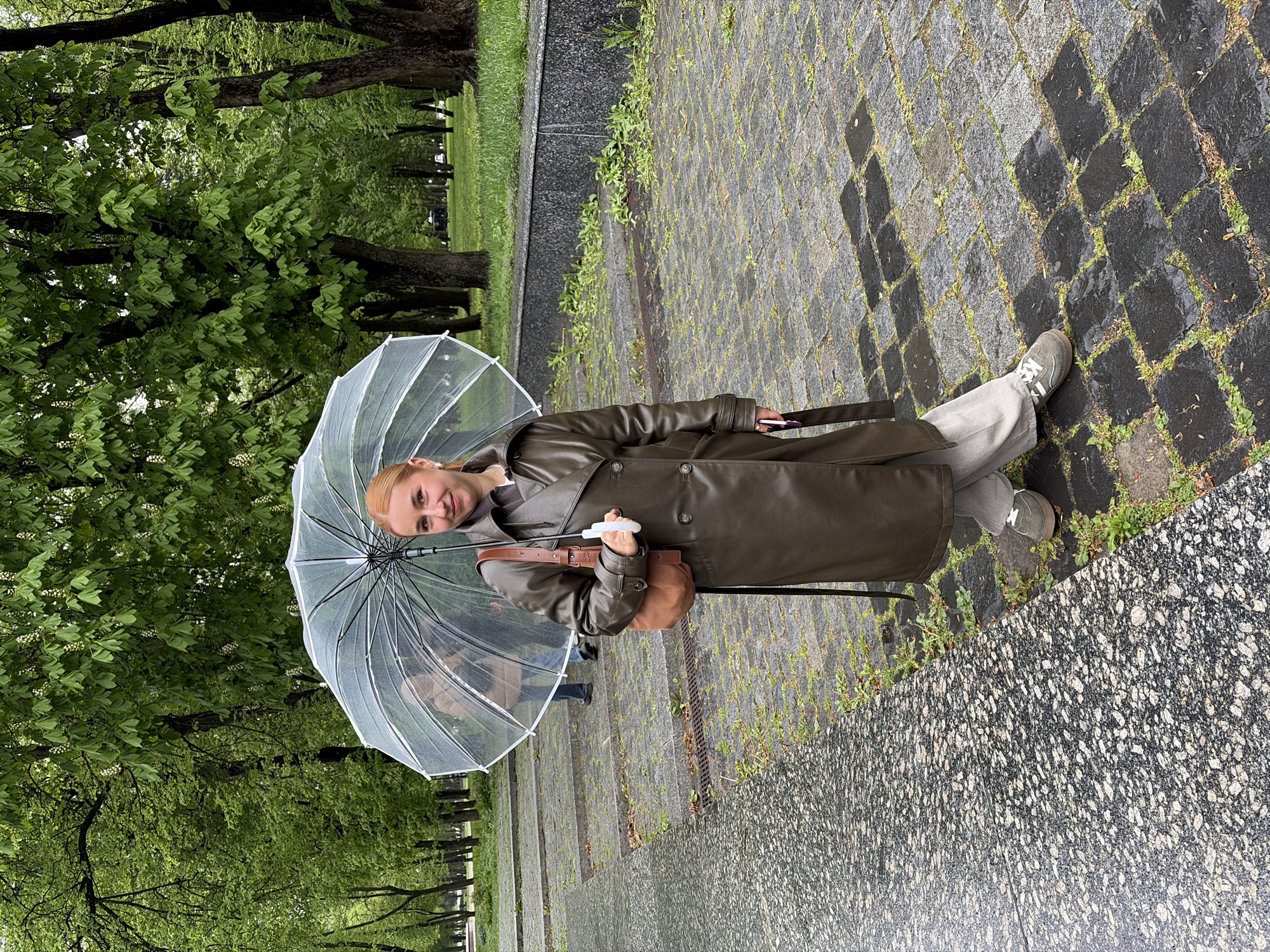 A woman stands on a damp, cobblestone path in a city park, holding a clear umbrella. She wears a long, olive-green trench coat, gray trousers, and gray sneakers. A brown shoulder bag hangs on her left side. Trees with green foliage surround the path.